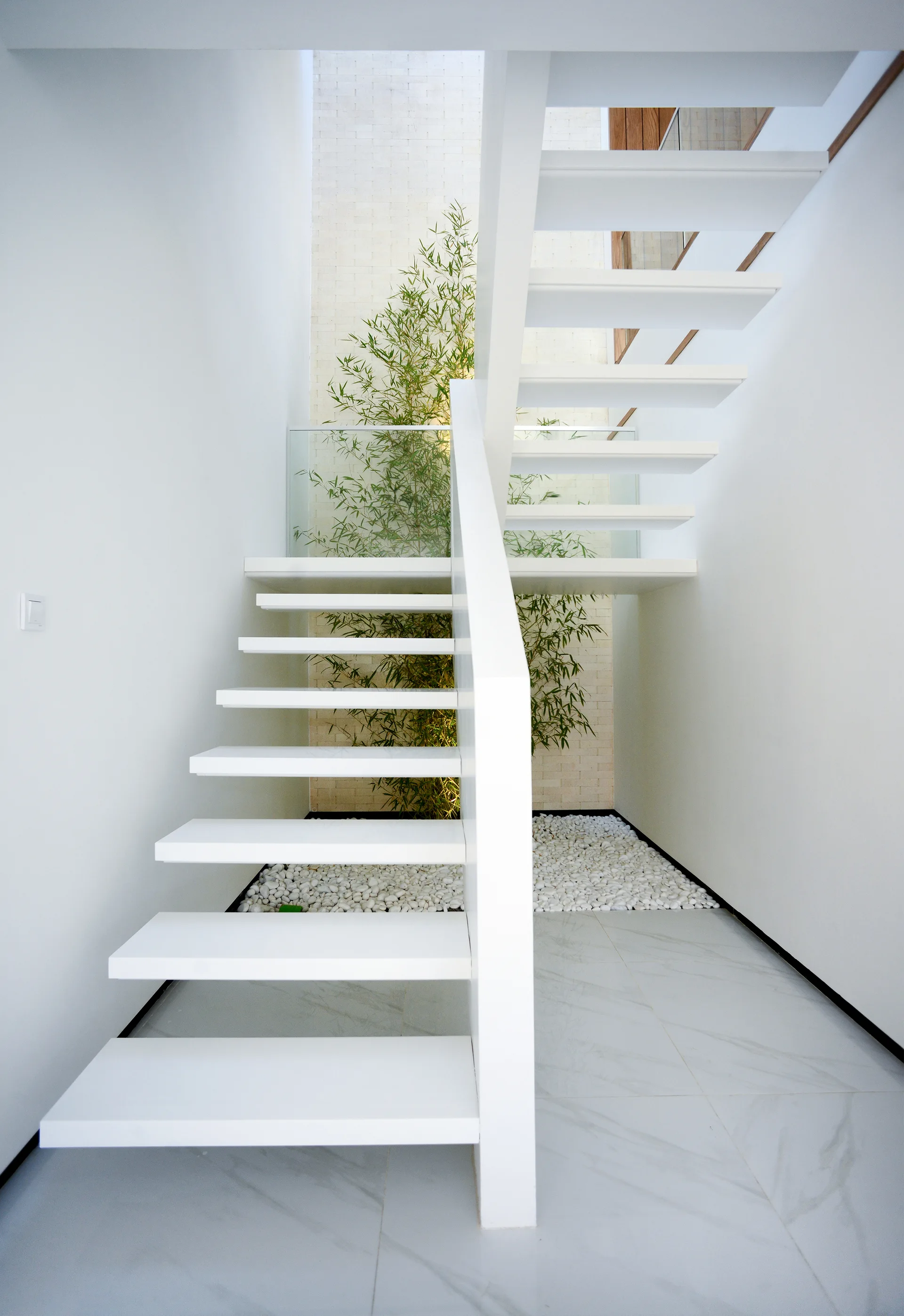 A modern white floating staircase rises above an indoor garden bed filled with white pebbles and a tall green bamboo plant against a textured stone wall.