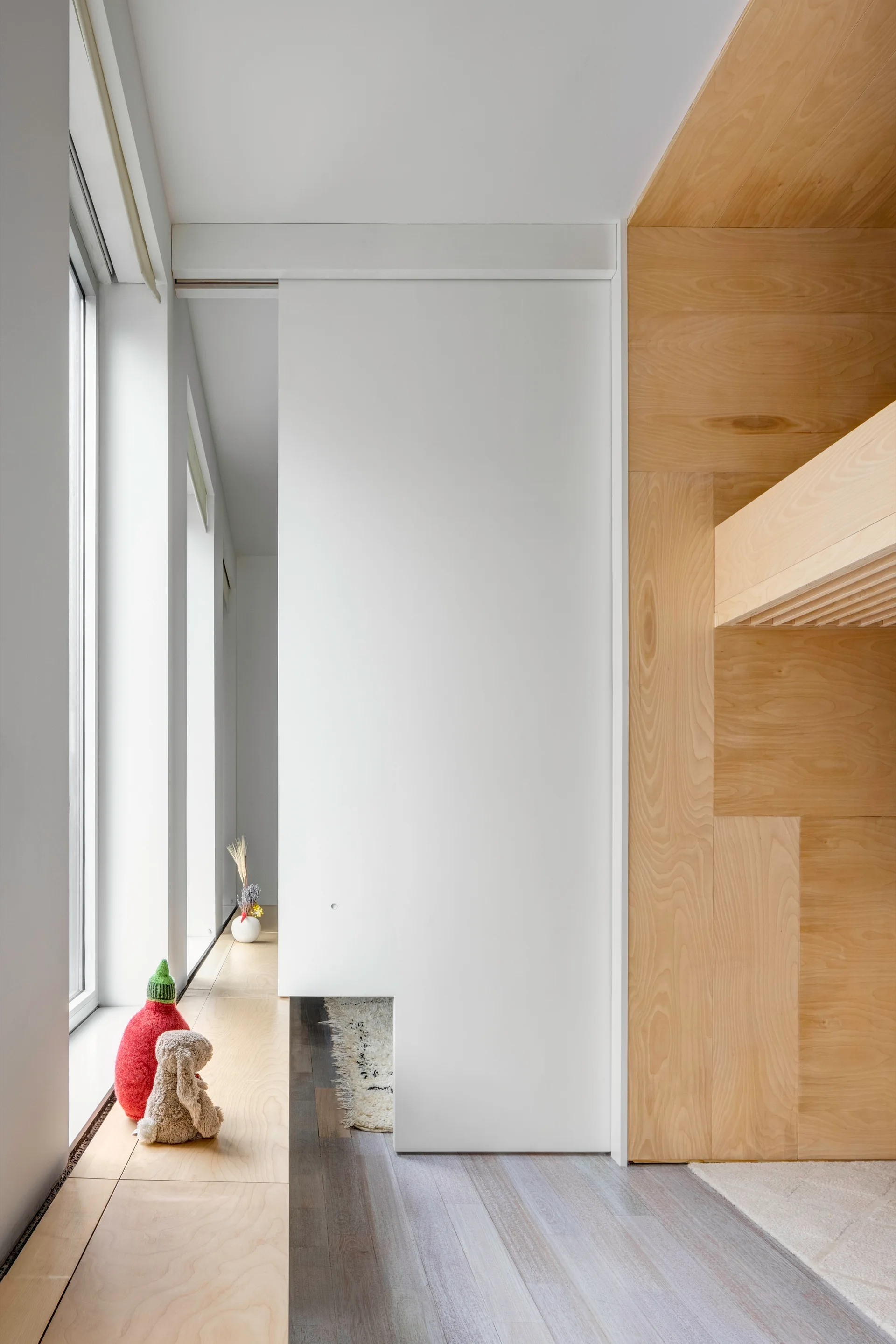 A minimalist interior featuring a white sliding partition wall, natural plywood millwork built-ins, and a matching wood sill bench beneath a window.