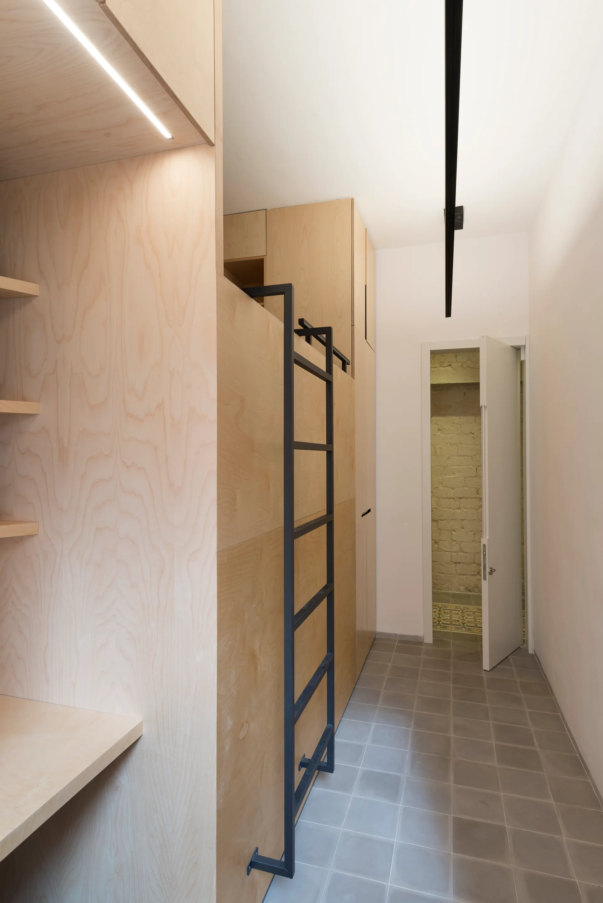 A minimalist hallway featuring tall plywood cabinets with an attached black metal ladder and square grey floor tiles leading to an open white door.