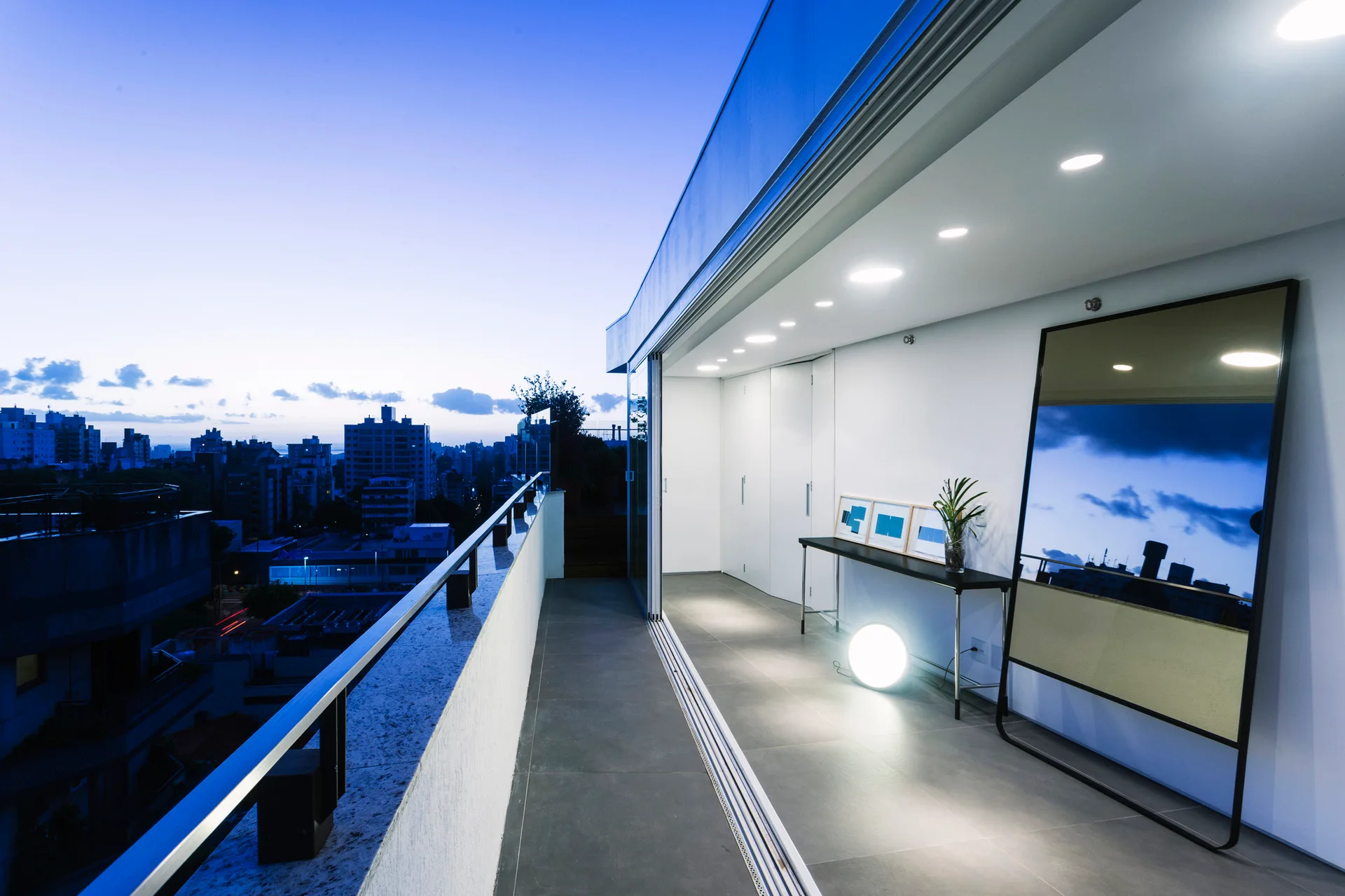 A modern balcony transition area at dusk with sliding glass doors, a large standing floor mirror, a slim console table, and recessed ceiling lights.