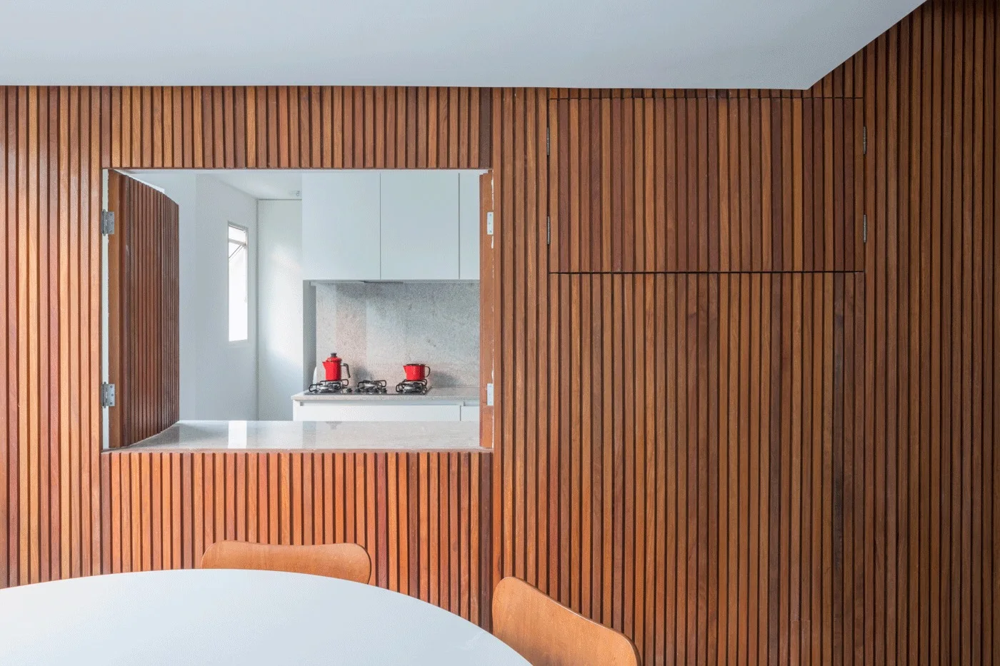 A vertical wood slat accent wall features a pass-through window opening to reveal a minimalist white kitchen with flat cabinets.