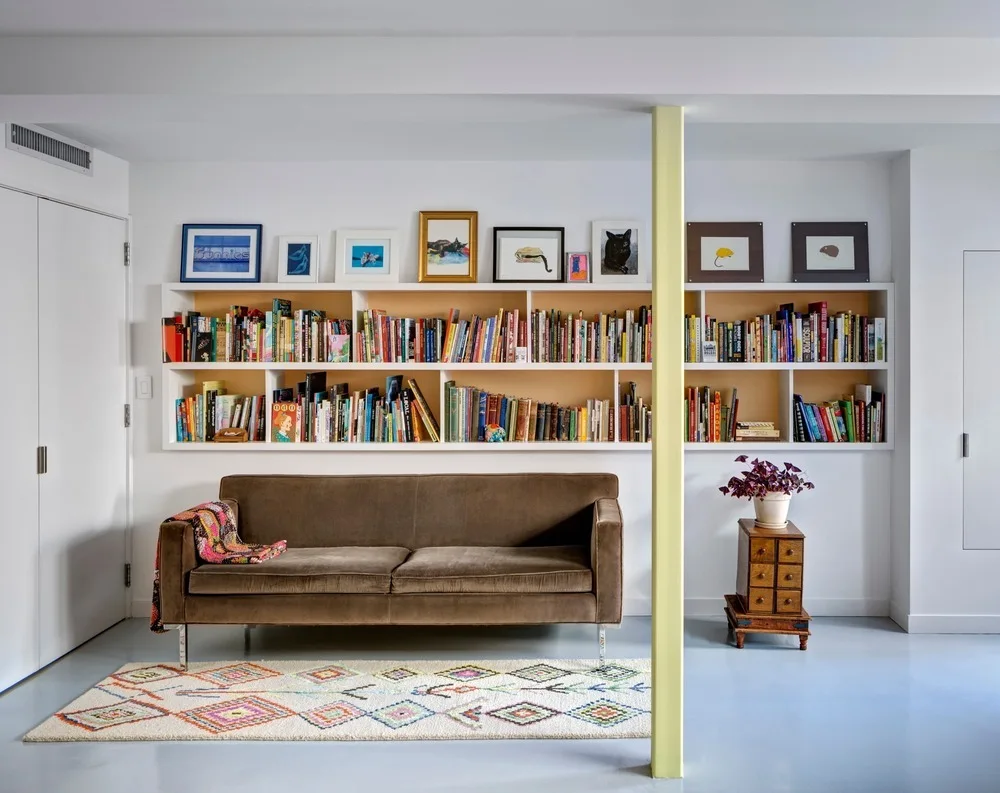 A brown velvet sofa sits before a long built-in bookshelf, next to a pale yellow support column on a light-colored floor.