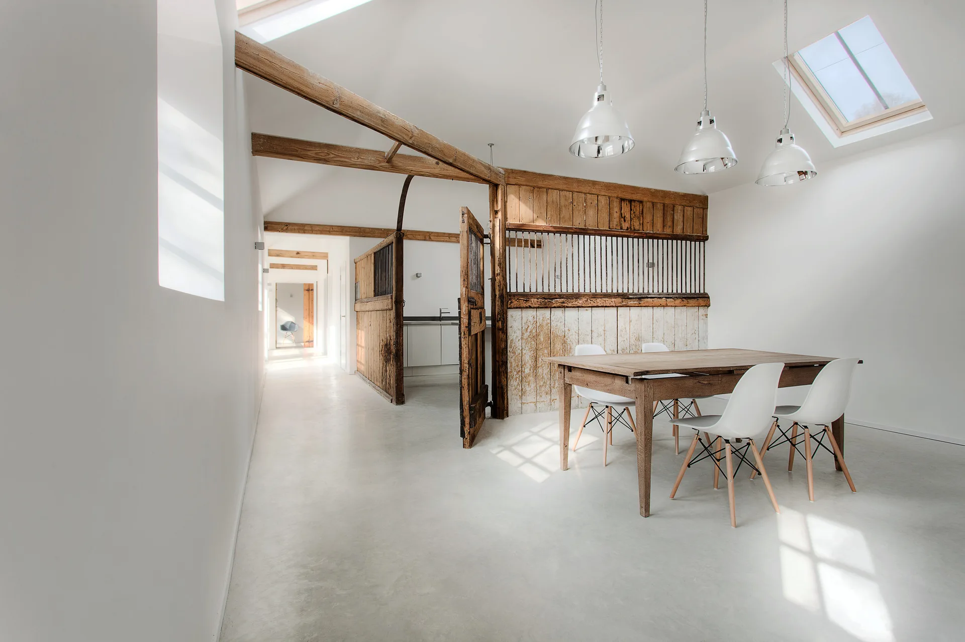 A light-filled dining room in a converted stable featuring a rustic wooden partition, a wooden table with white chairs, and polished concrete floors.