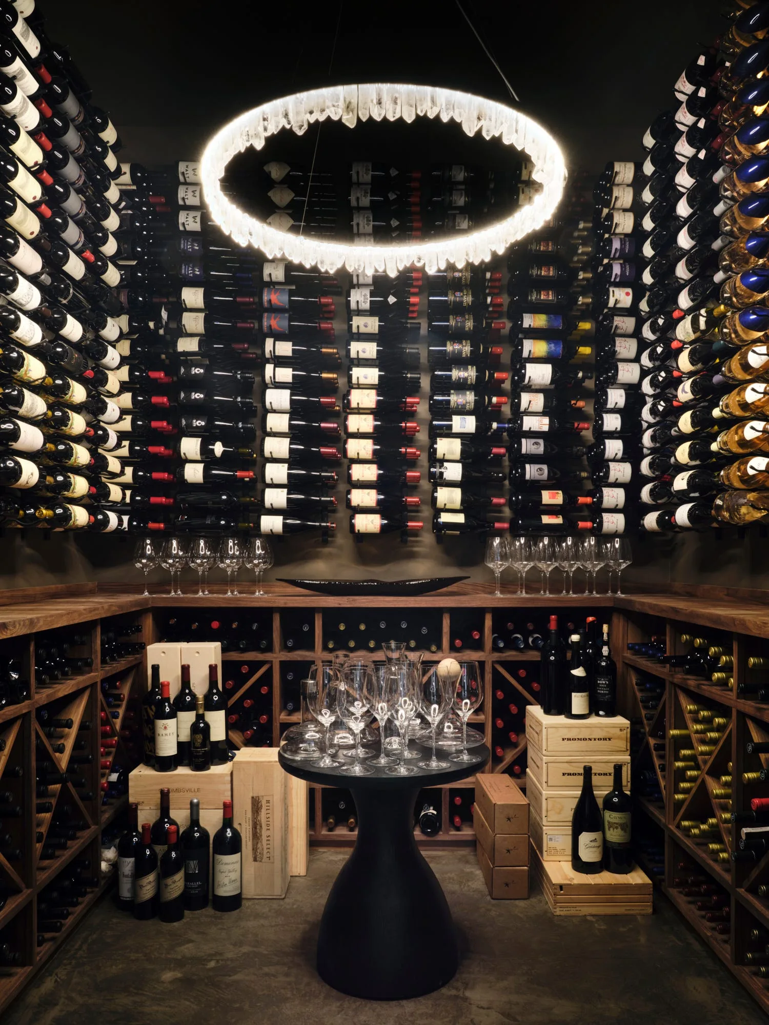 A dark wood wine cellar featuring floor-to-ceiling bottle racks, a central black pedestal table with wine glasses, and a circular crystal chandelier.