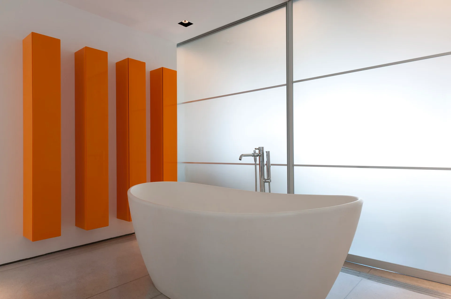 A white freestanding oval bathtub sits before a large frosted glass partition next to four vertical bright orange storage cabinets.