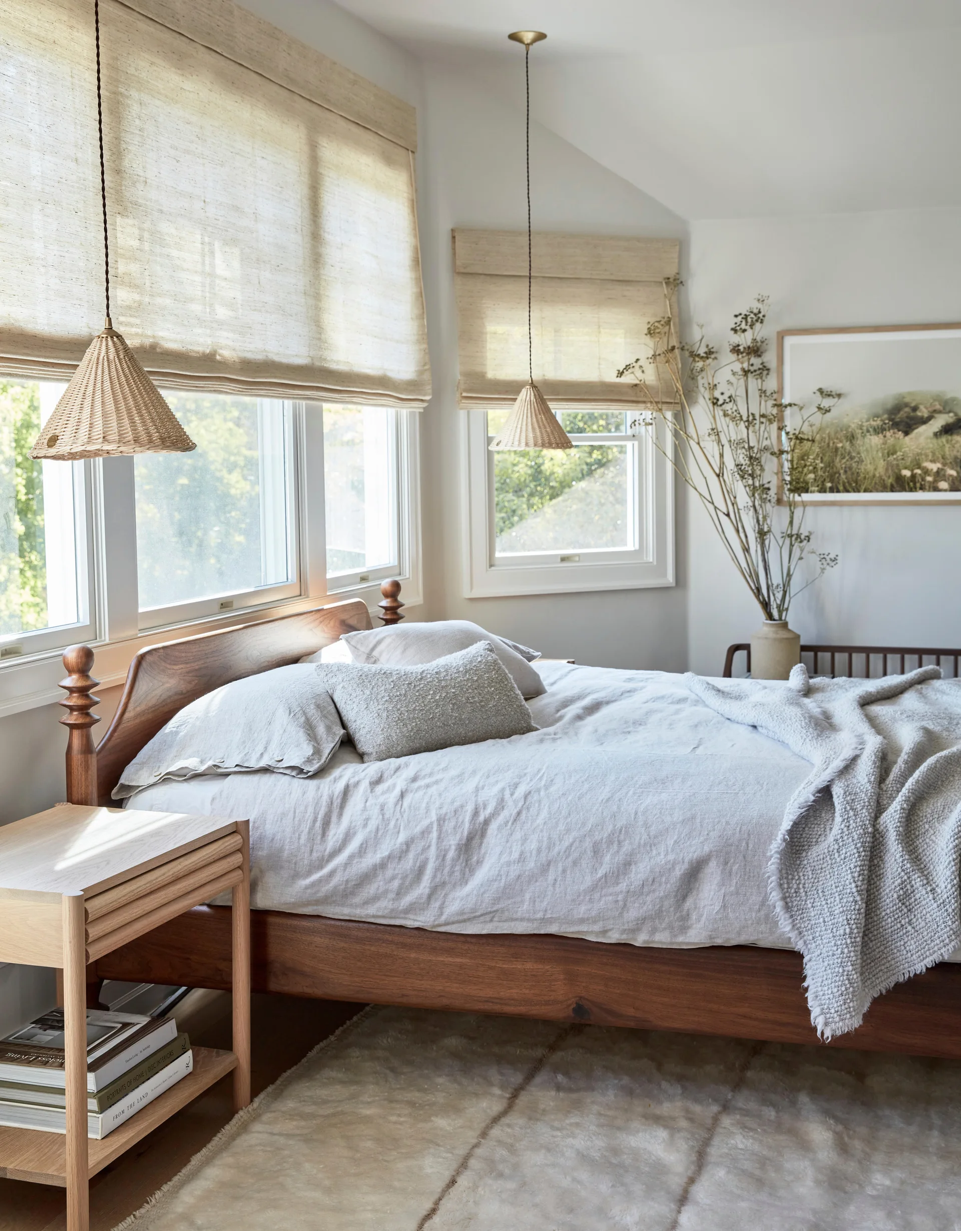 A tranquil bedroom features a dark wood bed frame, a light oak nightstand, and woven cone pendant lights hanging in front of windows with roman shades.