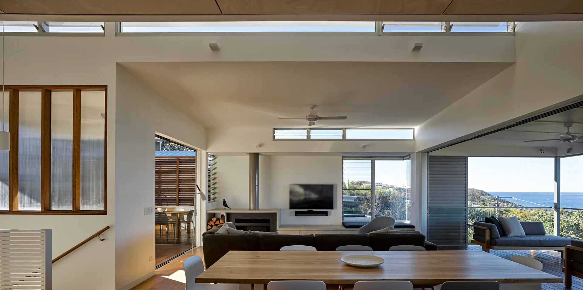 An open-plan coastal living and dining room with a wooden table, gray sectional sofa, fireplace, and large sliding doors opening to an ocean view.