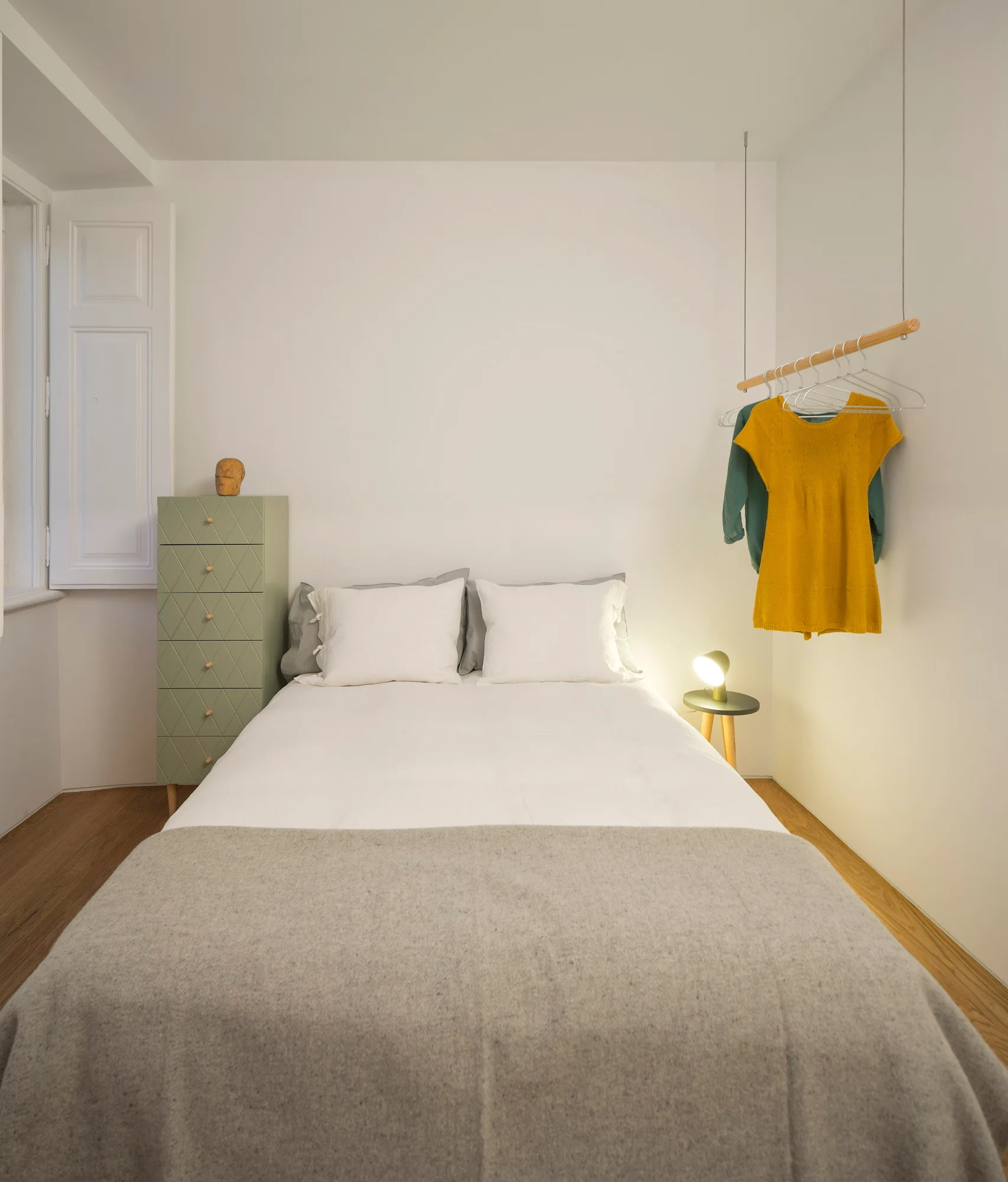 A minimalist bedroom features a bed with white and grey bedding next to a green tall chest and a suspended wooden clothing rod.