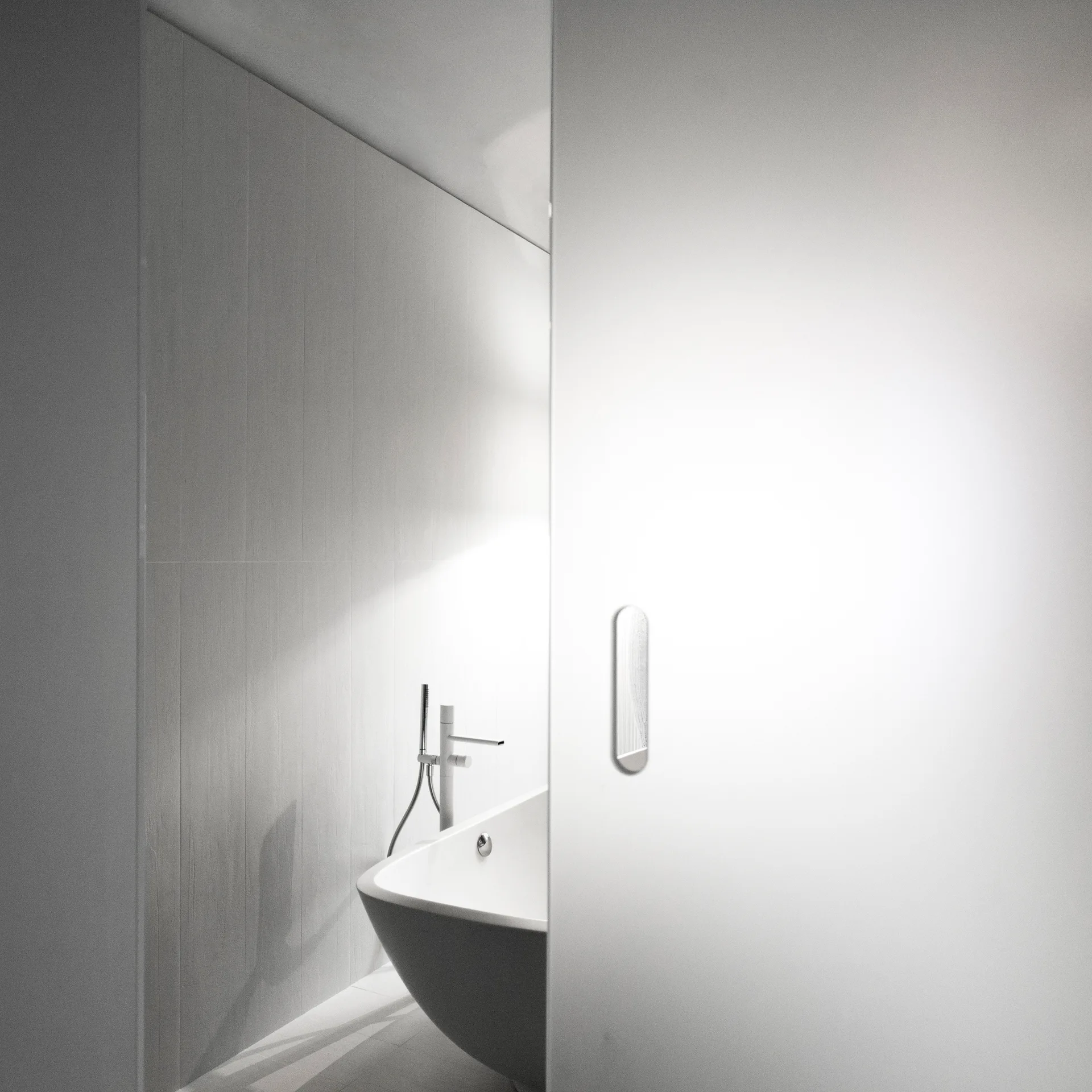 A modern white freestanding bathtub and chrome faucet set against a vertically paneled wall, partially obscured by a minimalist sliding door.
