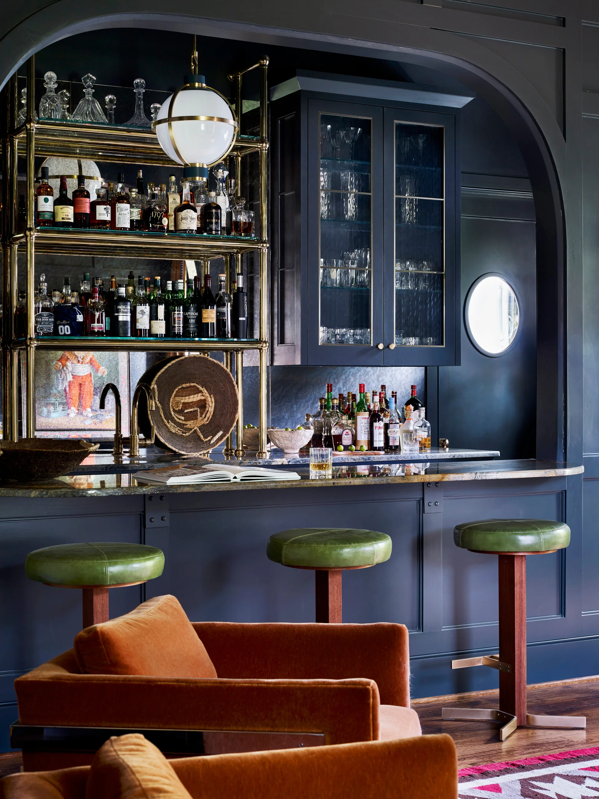 A home bar with dark blue wall paneling, a brass bottle rack, three green stools, and orange velvet armchairs in the foreground.