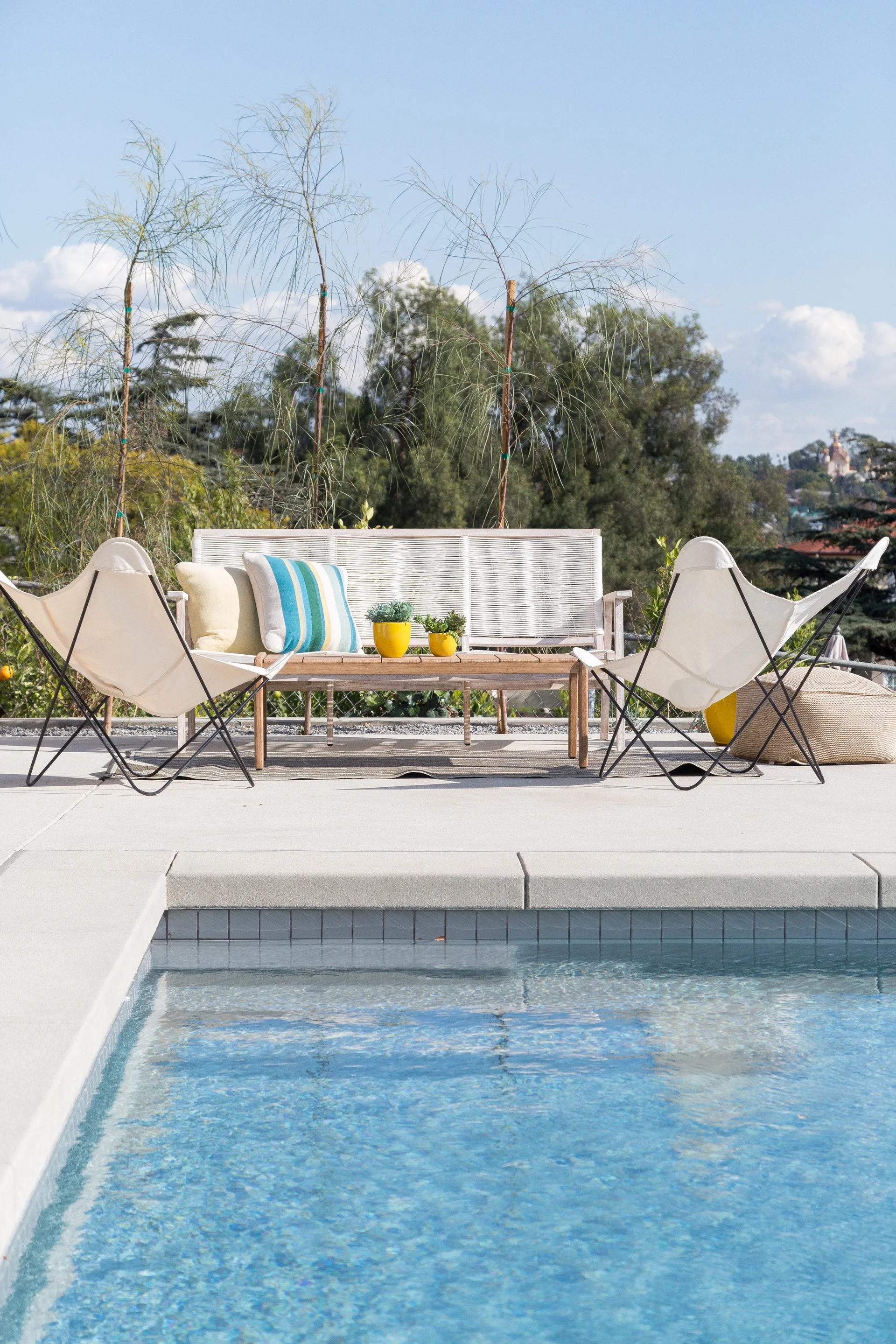 Modern poolside patio furniture including a woven rope sofa and two white butterfly chairs arranged on a concrete deck next to a swimming pool.