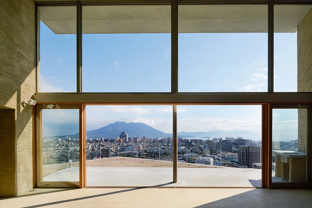 A minimalist room features a large floor-to-ceiling panoramic window looking out onto a city and a mountain, framed by textured rammed earth walls.