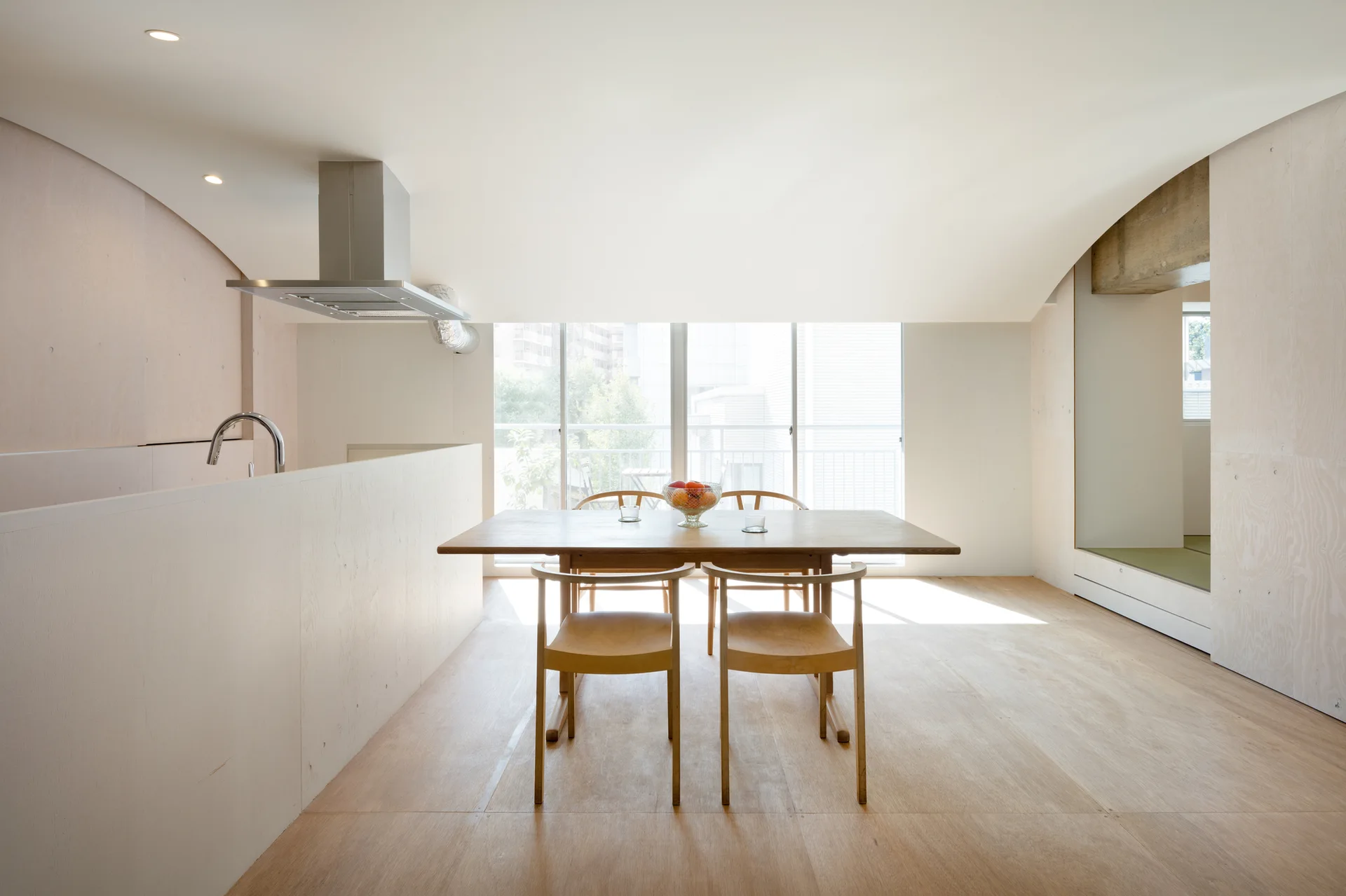 A wooden dining set sits in a minimalist open-plan kitchen with pale plywood walls, a curved white ceiling, and a large window.