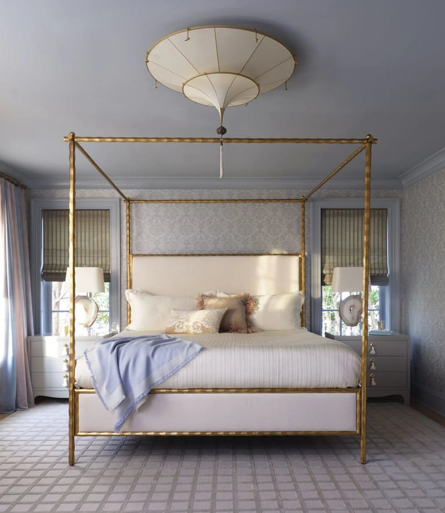 A gold four-poster canopy bed sits centered in a bedroom with dusty blue walls, patterned wallpaper, grey nightstands, and a unique pendant light.