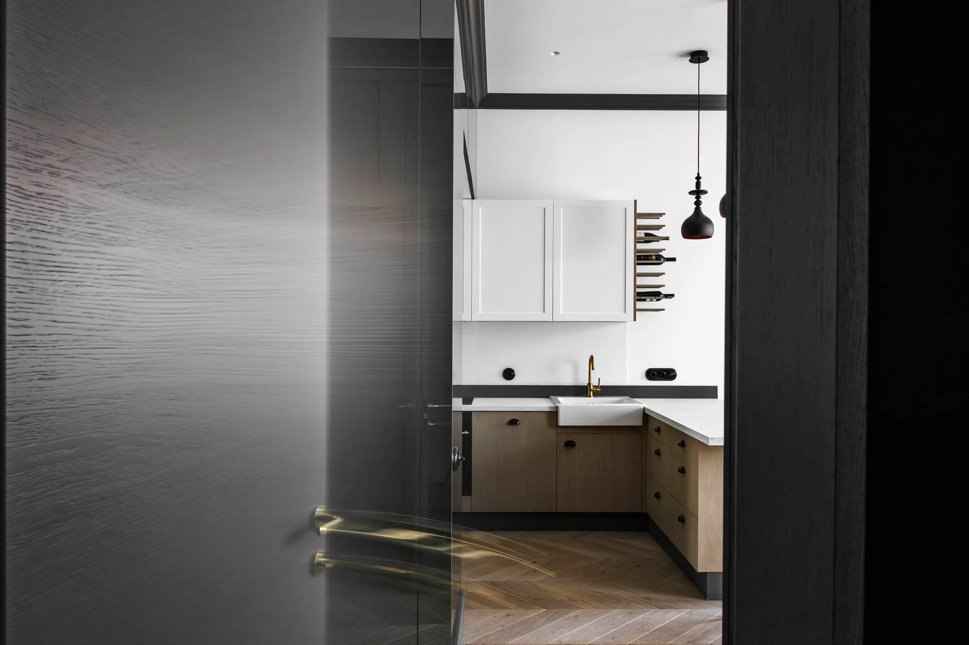 A modern kitchen viewed through a doorway, featuring wood lower cabinets, white upper cabinets, a gold faucet, and herringbone wood flooring.