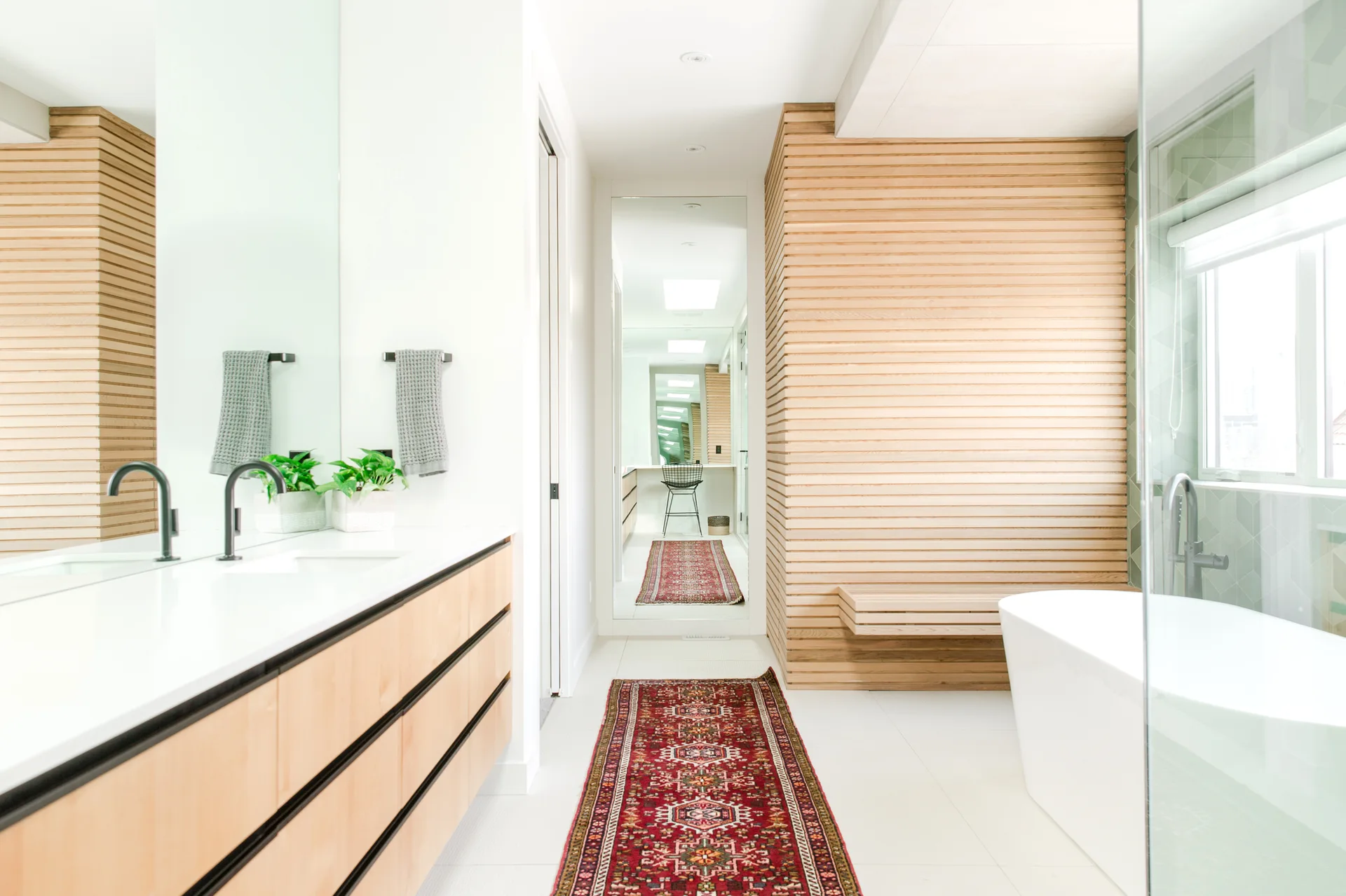 A contemporary bathroom features a light wood vanity, a white freestanding tub, and a red patterned runner rug on the white tiled floor.
