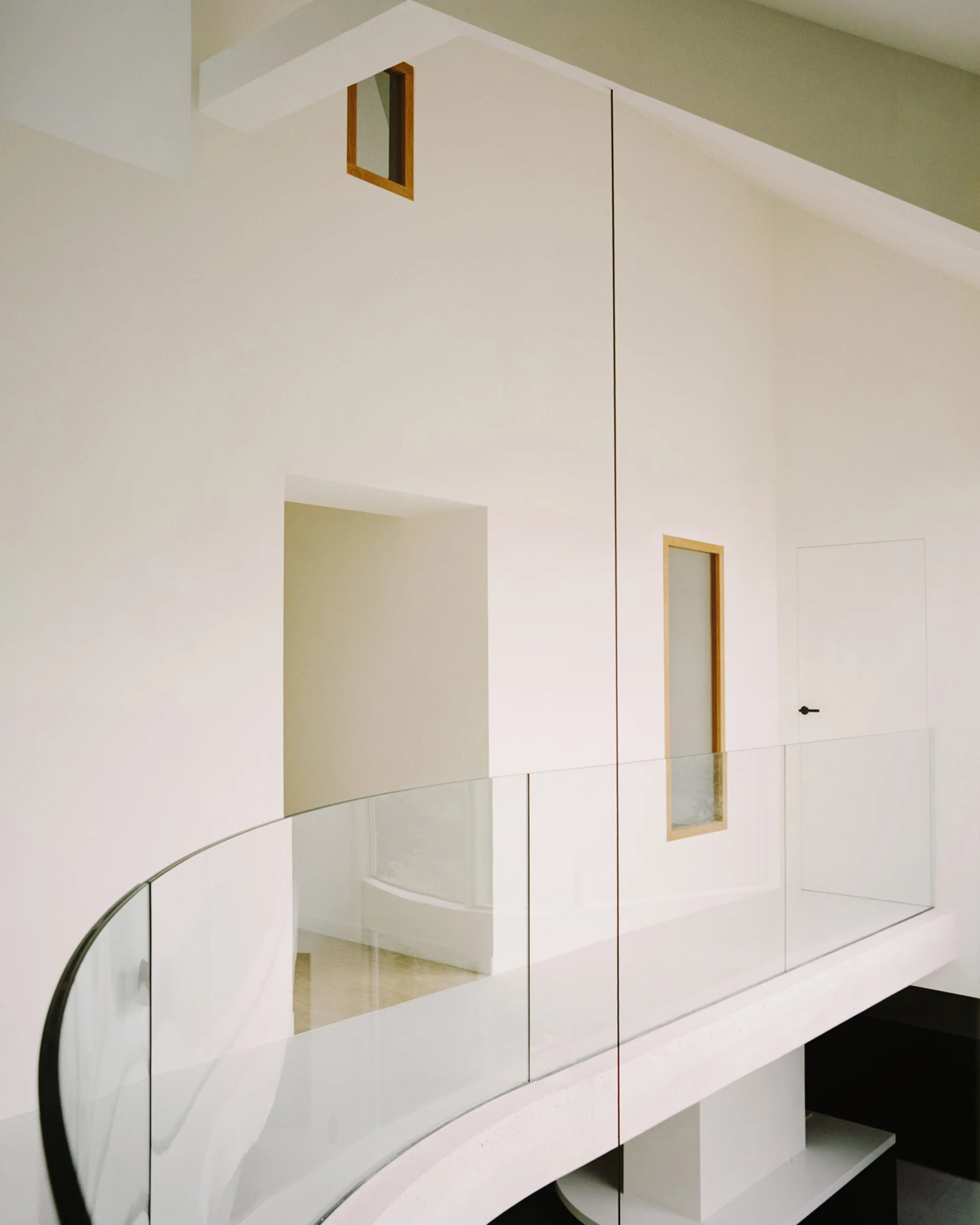 A minimalist residential landing featuring a curved glass balustrade railing alongside smooth off-white plaster walls with small wooden-framed interior windows.
