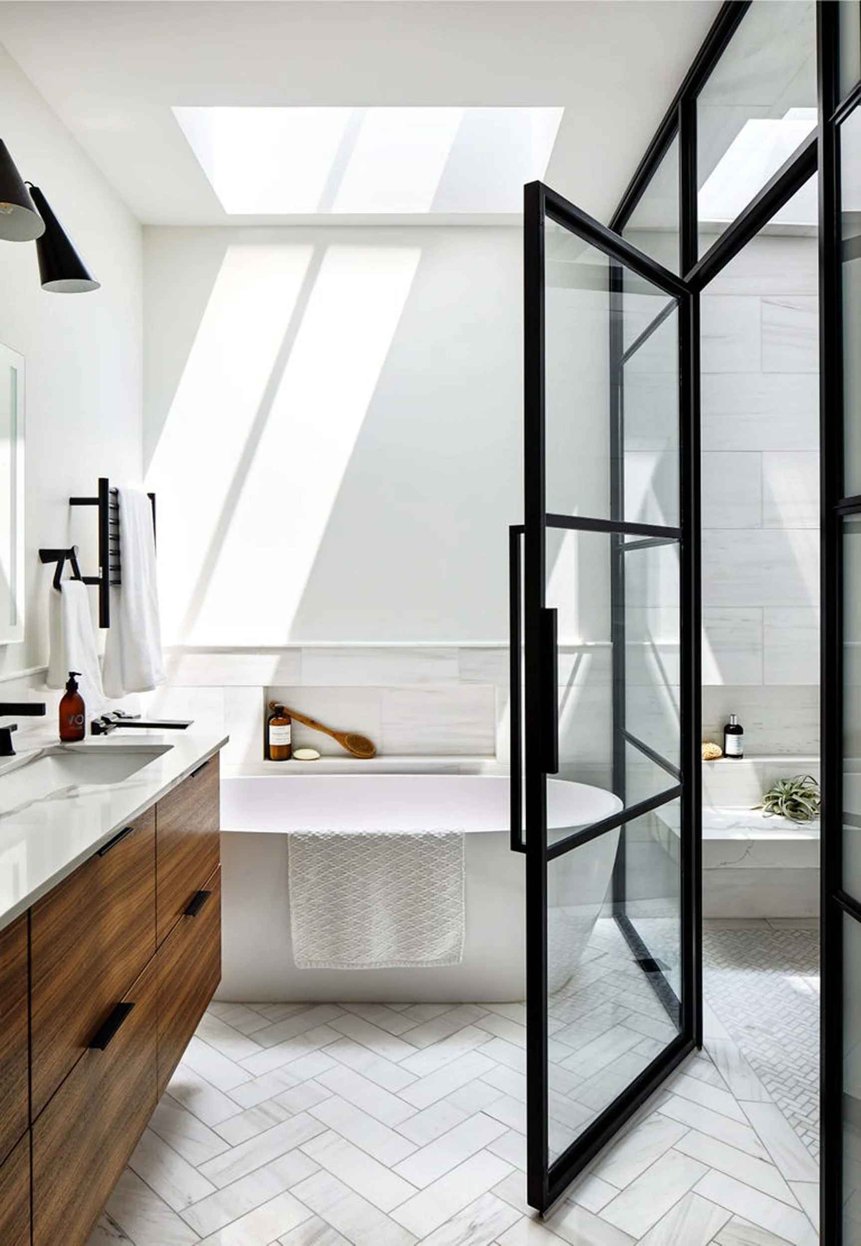 A modern bathroom with a wood vanity, white soaking tub, and black metal framed glass shower door illuminated by a large skylight.