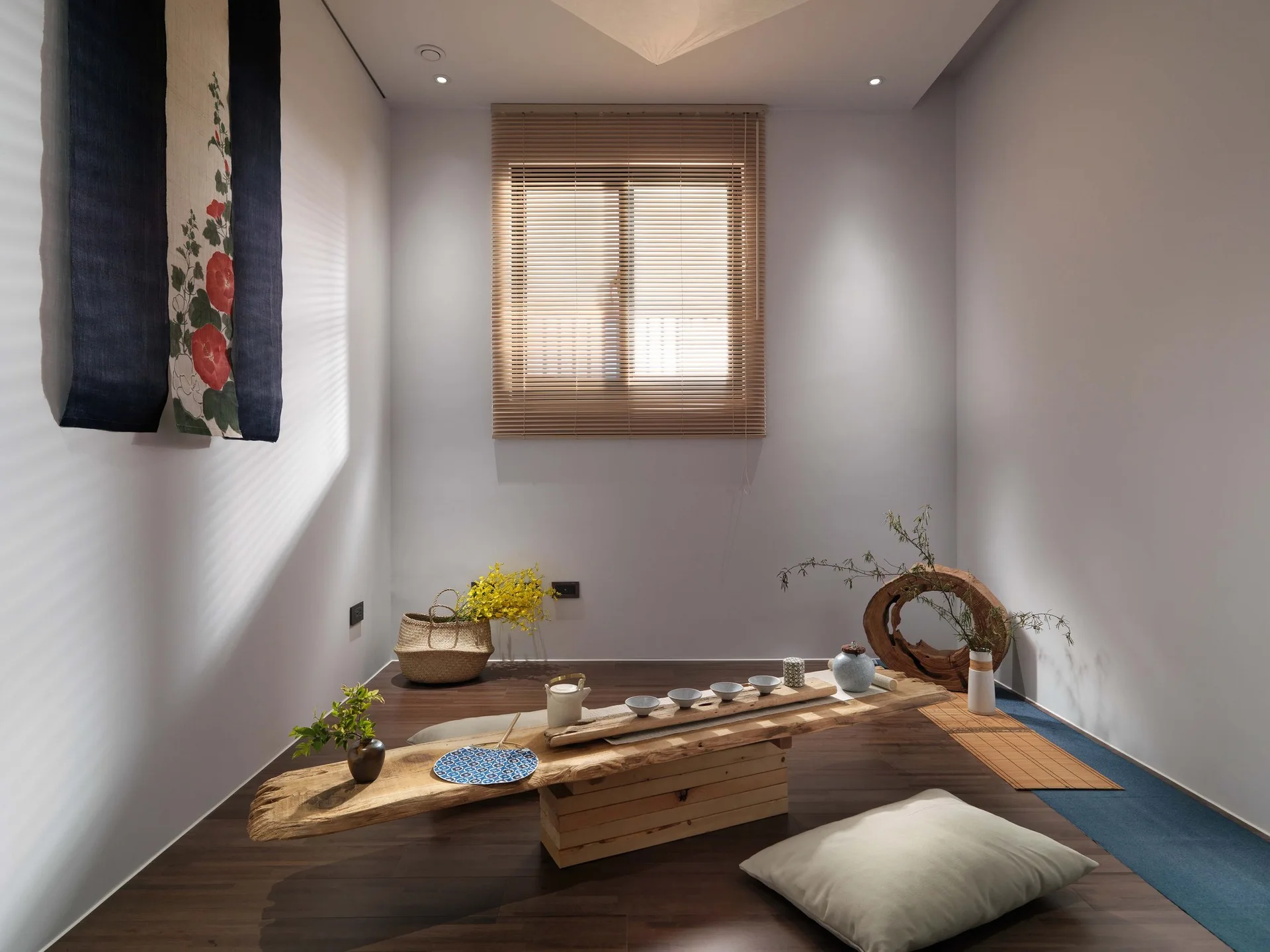 A minimalist tea room features a rustic low wooden slab table, beige floor cushions, and wooden window blinds on a dark wood floor.