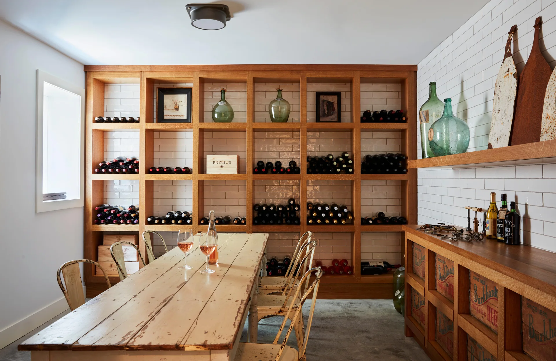 A rustic wine tasting room featuring a distressed wooden table, vintage metal chairs, a white subway tile wall, and large wood grid wine shelving.