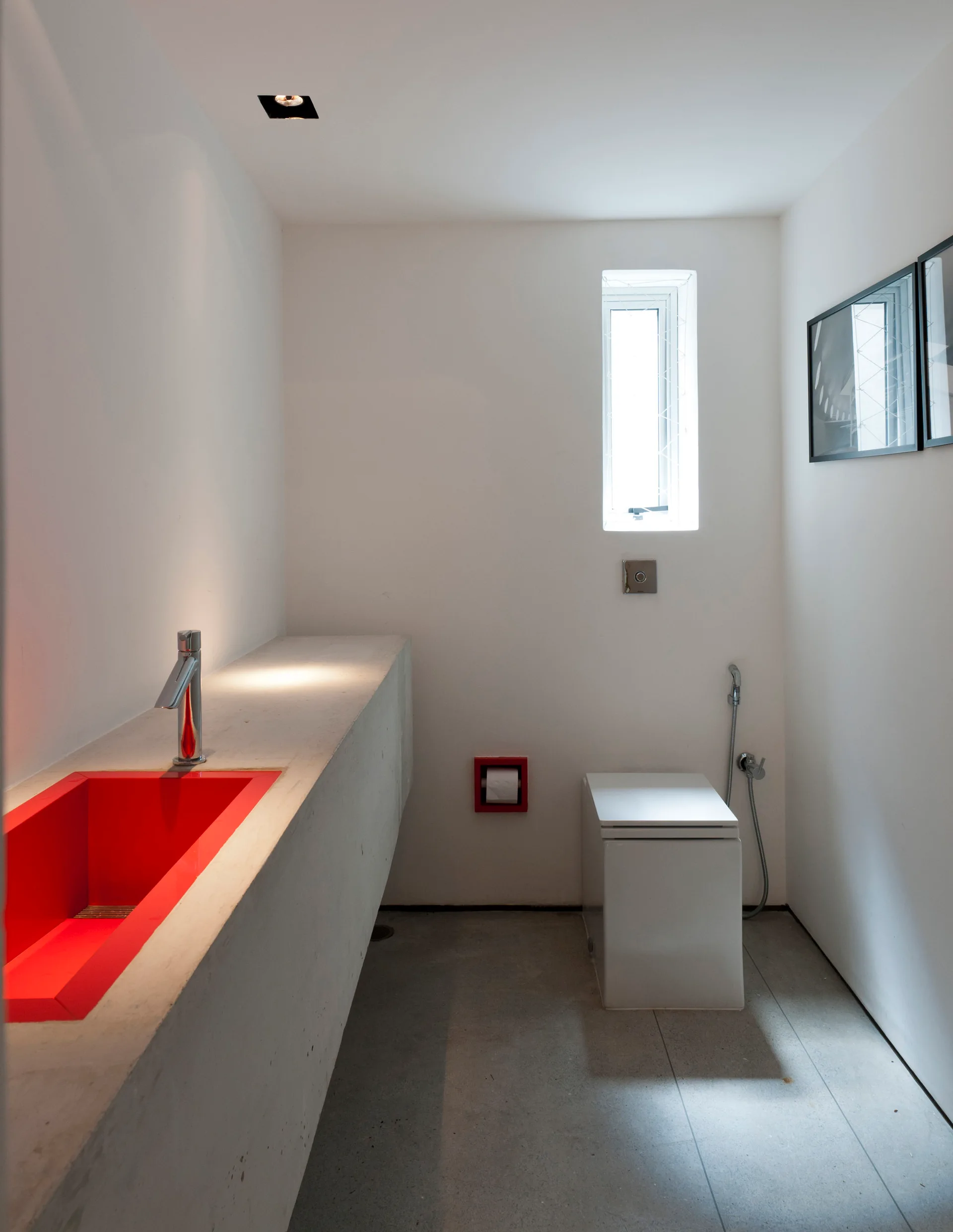 A minimalist powder room featuring a gray concrete vanity with an inset bright red sink, a white wall-mounted toilet, and plain white walls.