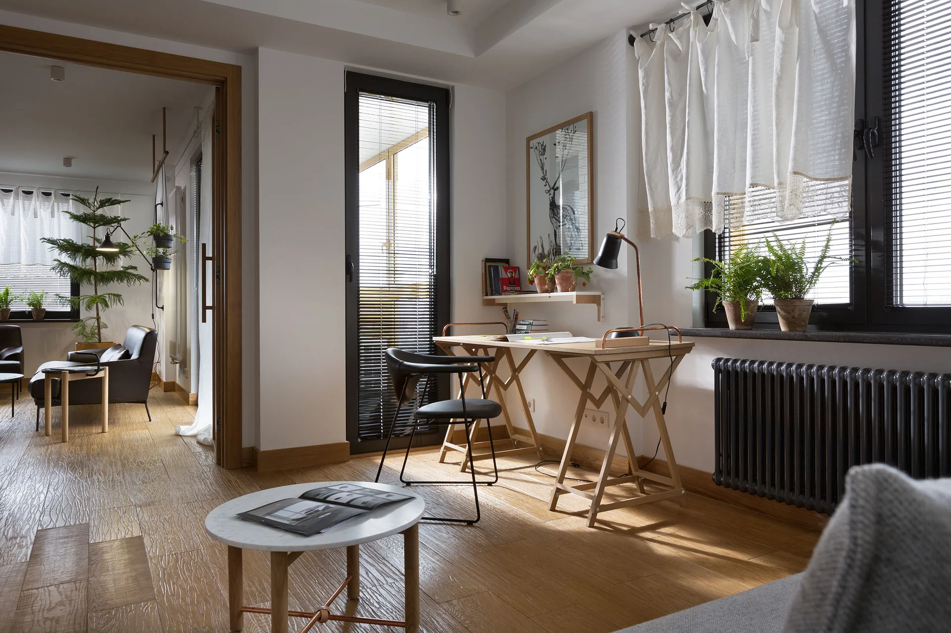 A home office corner features a wooden x-frame desk, a black task chair, potted plants on the windowsill, and a dark grey radiator.