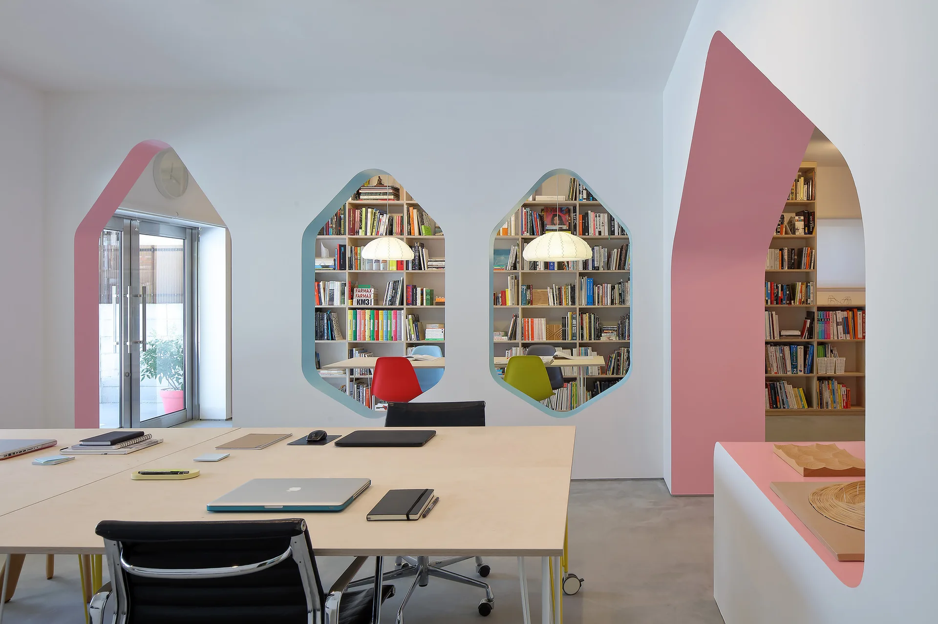A modern white office features a light wood table, black chairs, geometric wall cutouts revealing bookshelves, and pink-painted arched doorways.