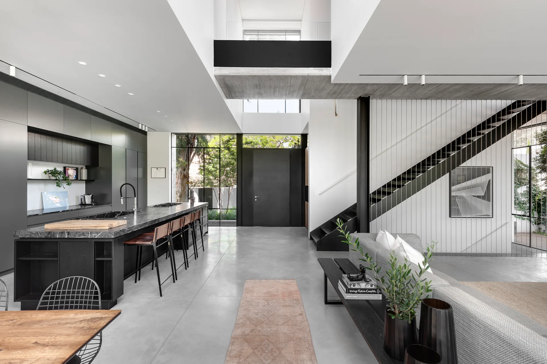 A spacious open-plan kitchen and living space featuring a black stone island, a black metal staircase, and smooth concrete flooring.