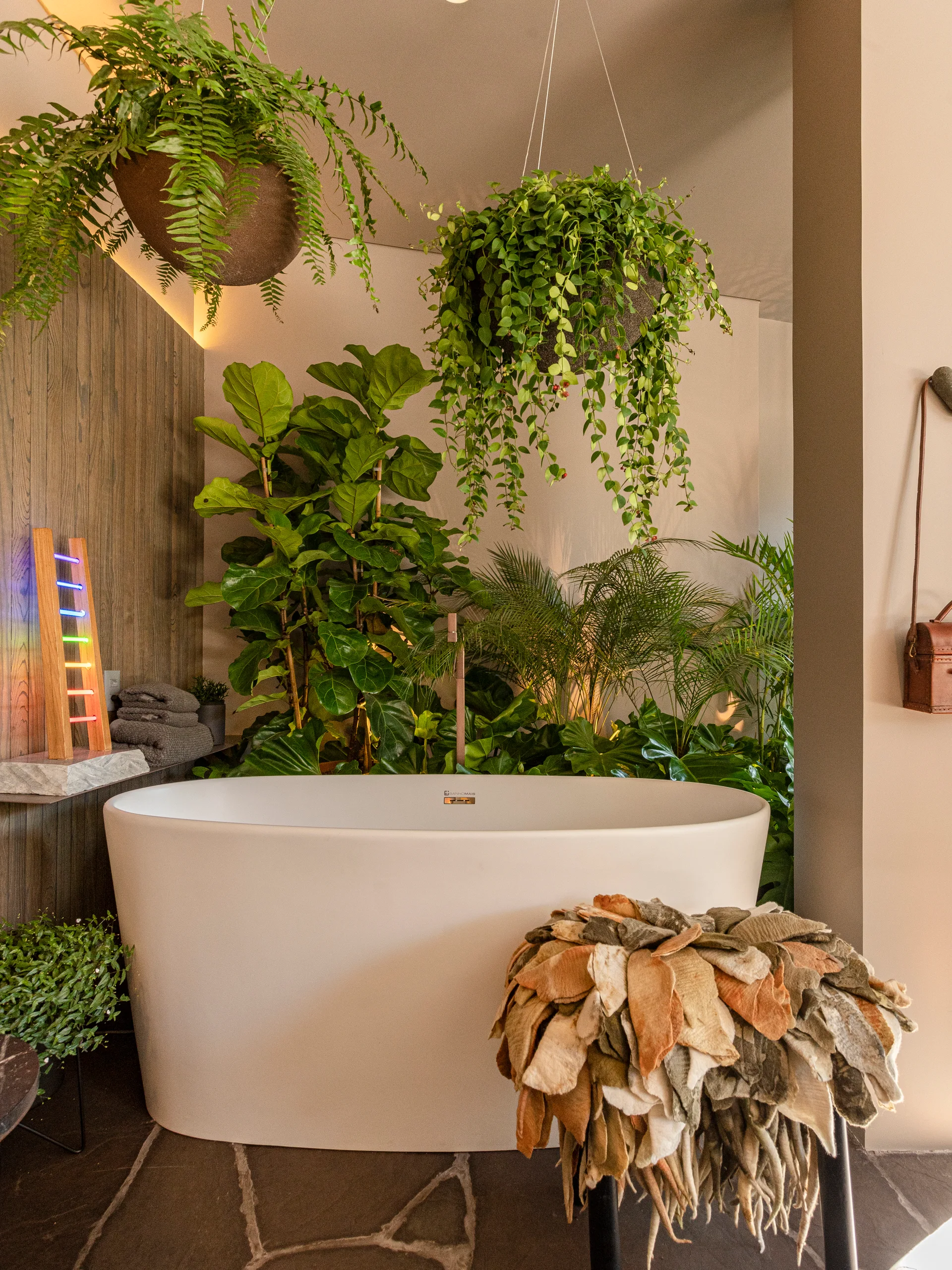 A white freestanding bathtub surrounded by dense green houseplants, including hanging ferns and a fiddle-leaf fig, against a wood-paneled wall.