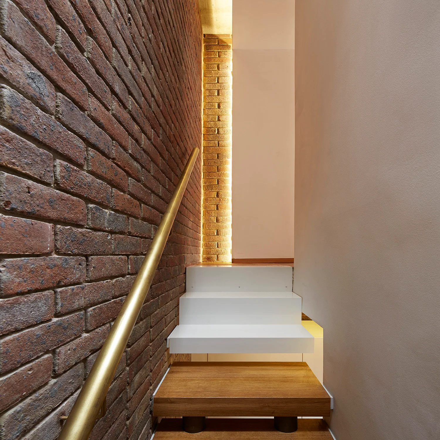 A modern staircase featuring a combination of wood and white floating treads next to an exposed brick wall with a gold handrail.