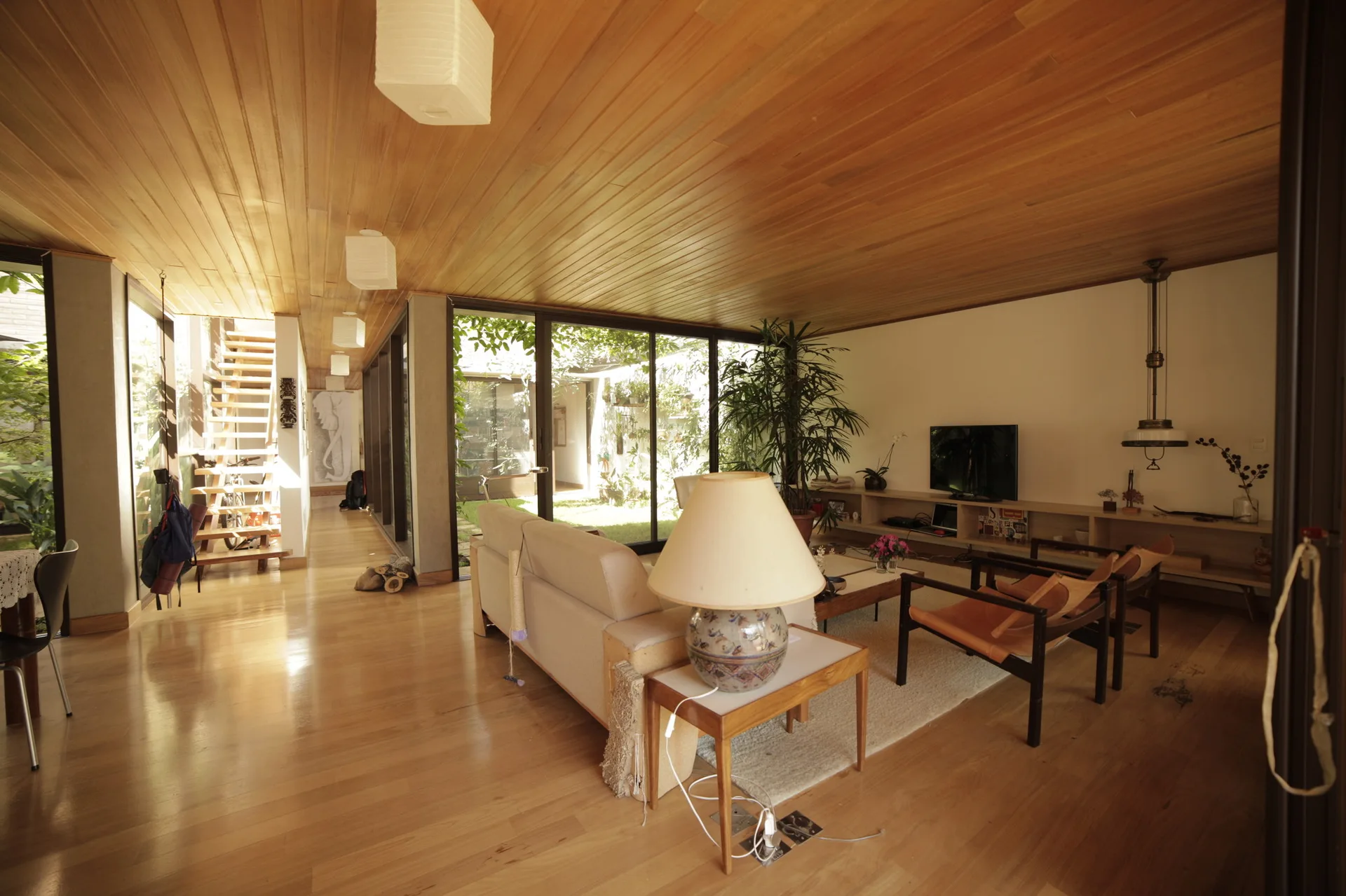 A mid-century modern living room features a warm wood-planked ceiling, polished hardwood floors, a beige sofa, and sliding glass doors looking onto a garden.