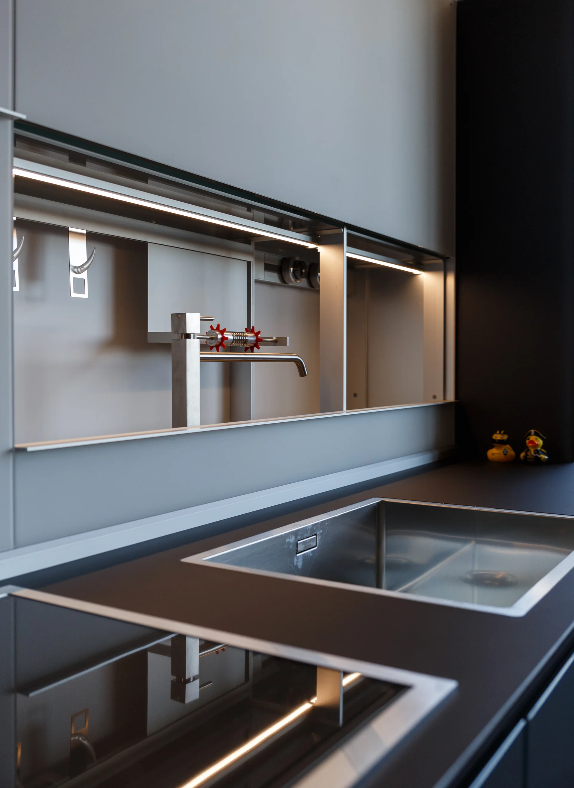A dark matte countertop holds a stainless steel sink beneath a grey recessed backsplash shelf featuring integrated LED strip lighting and a modern faucet.