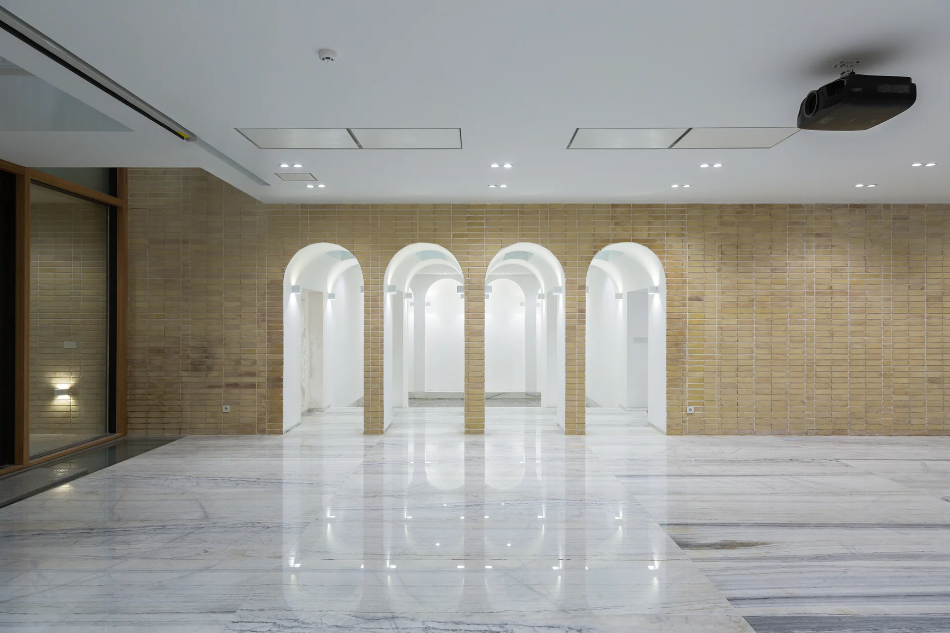 An expansive minimalist interior features a beige brick wall with four arched portals, a highly reflective polished stone floor, and a white ceiling.