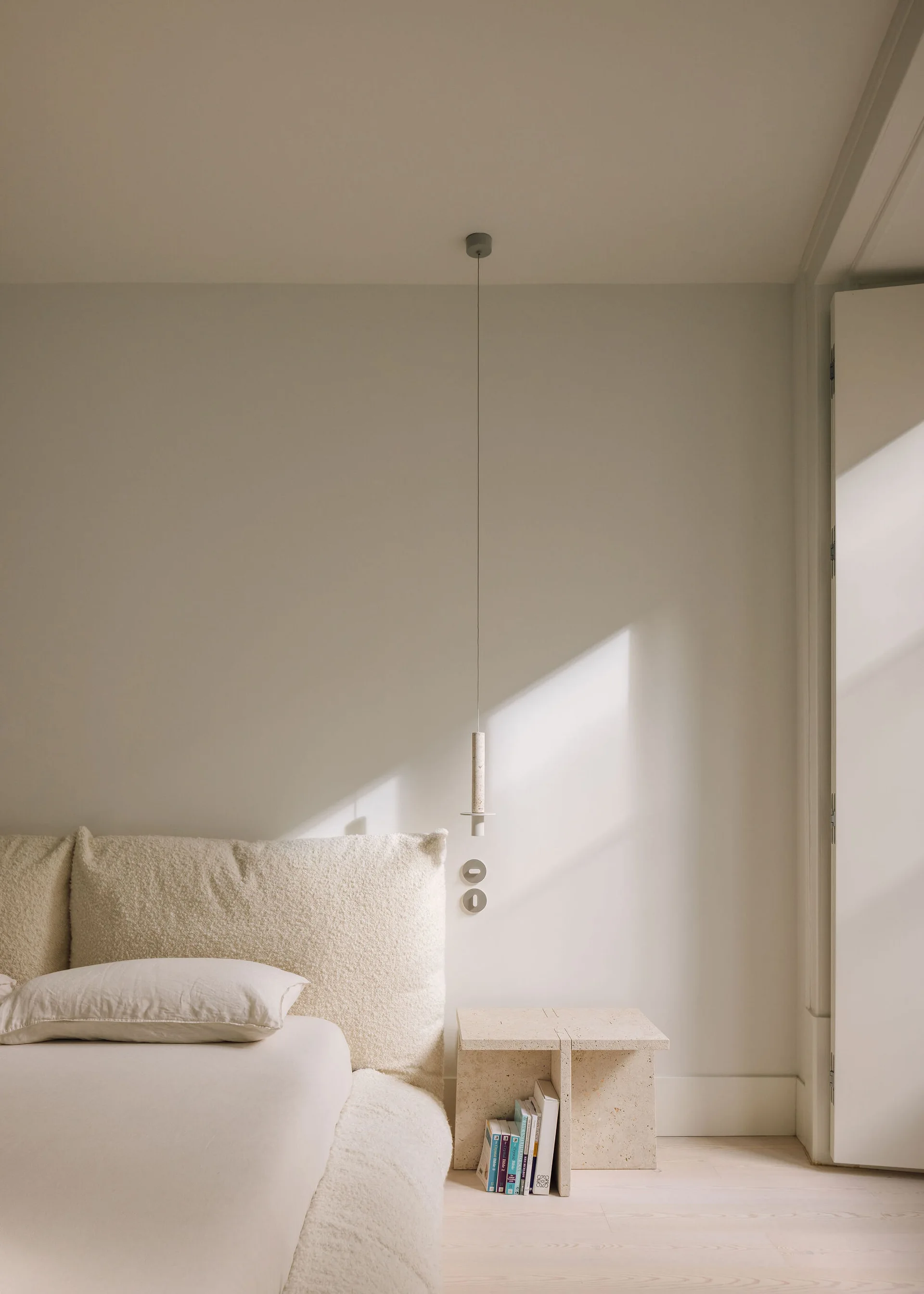 A minimalist bedroom features an off-white boucle bed, a travertine stone side table with books, and a slim pendant light hanging from the ceiling.