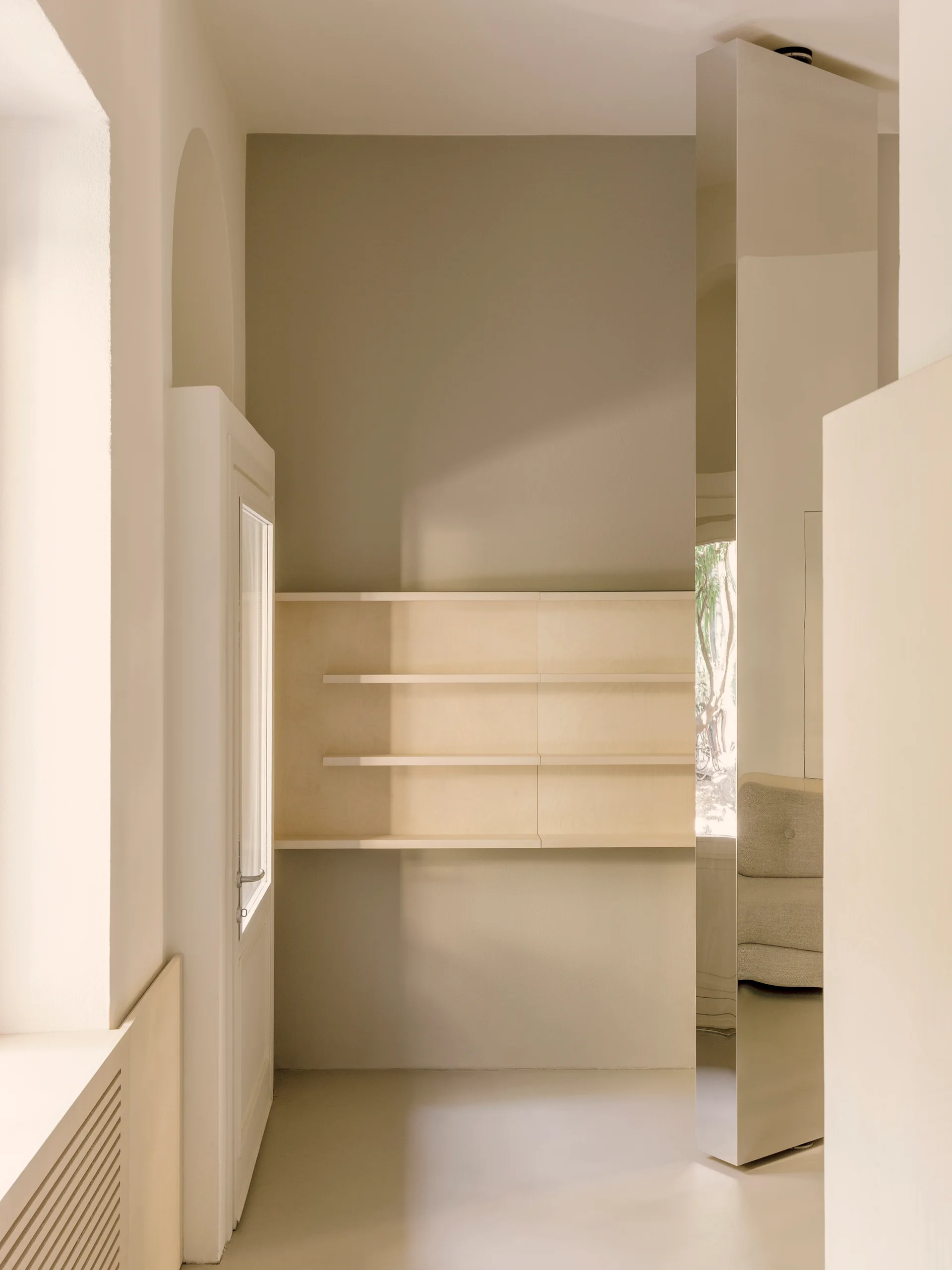 A minimalist beige hallway features light wood built-in shelving, a paneled door, and a tall floor-to-ceiling mirrored column.