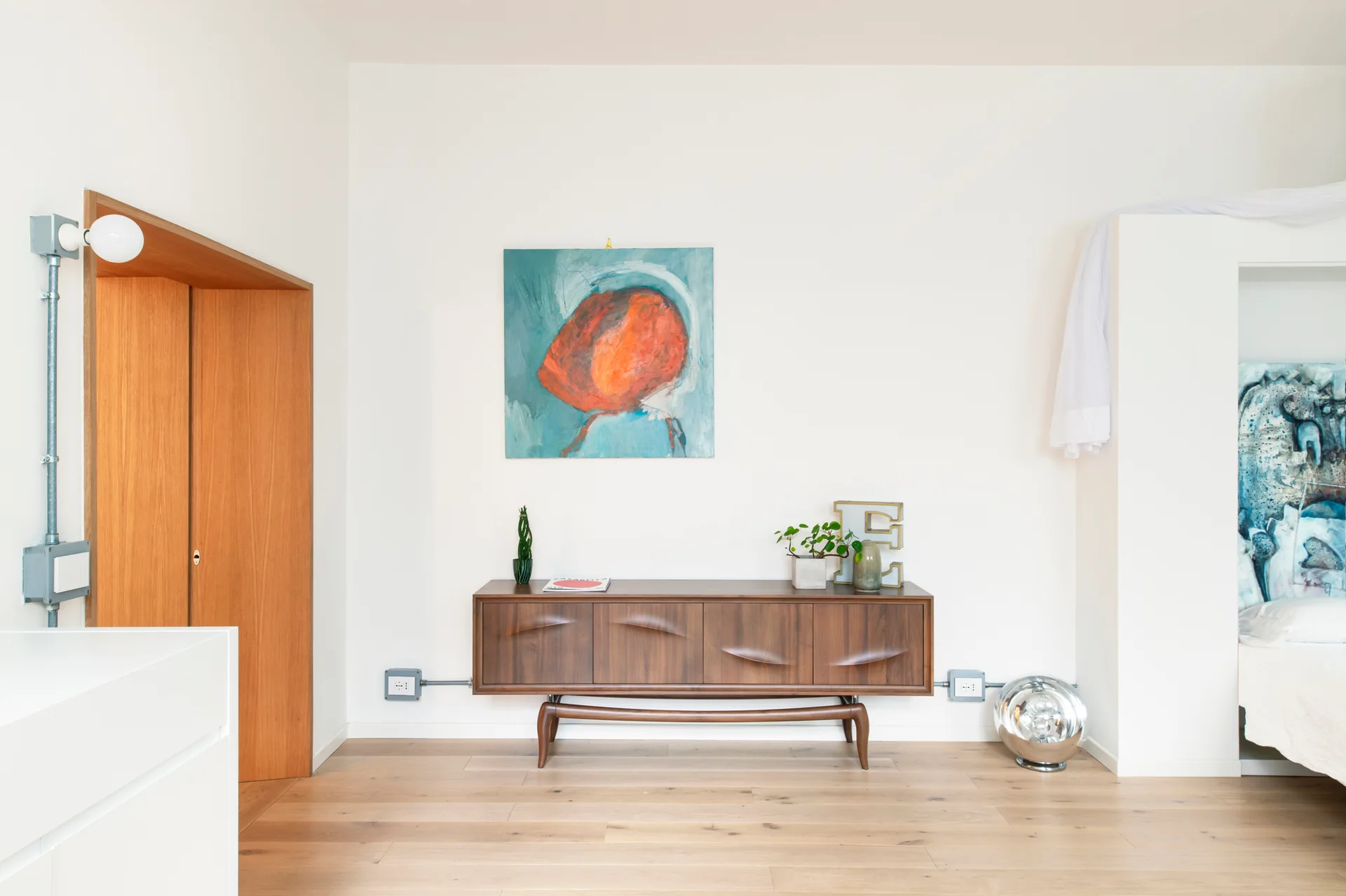 A minimalist bedroom featuring a mid-century walnut sideboard beneath an abstract painting, light wood floors, and a wood-lined doorway.