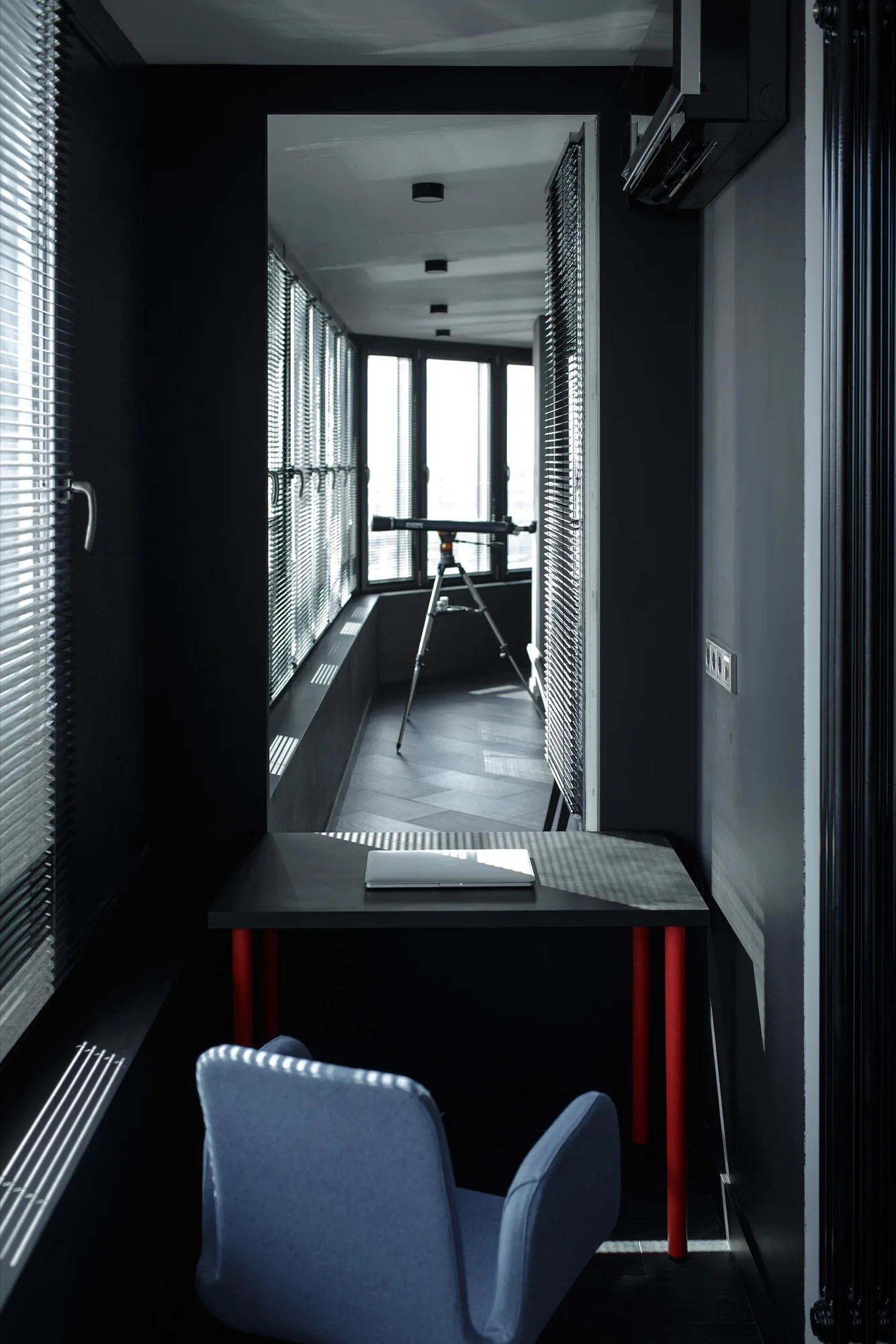 A dark home office features a desk with red legs, a blue armchair, and a view into a bright balcony containing a telescope.