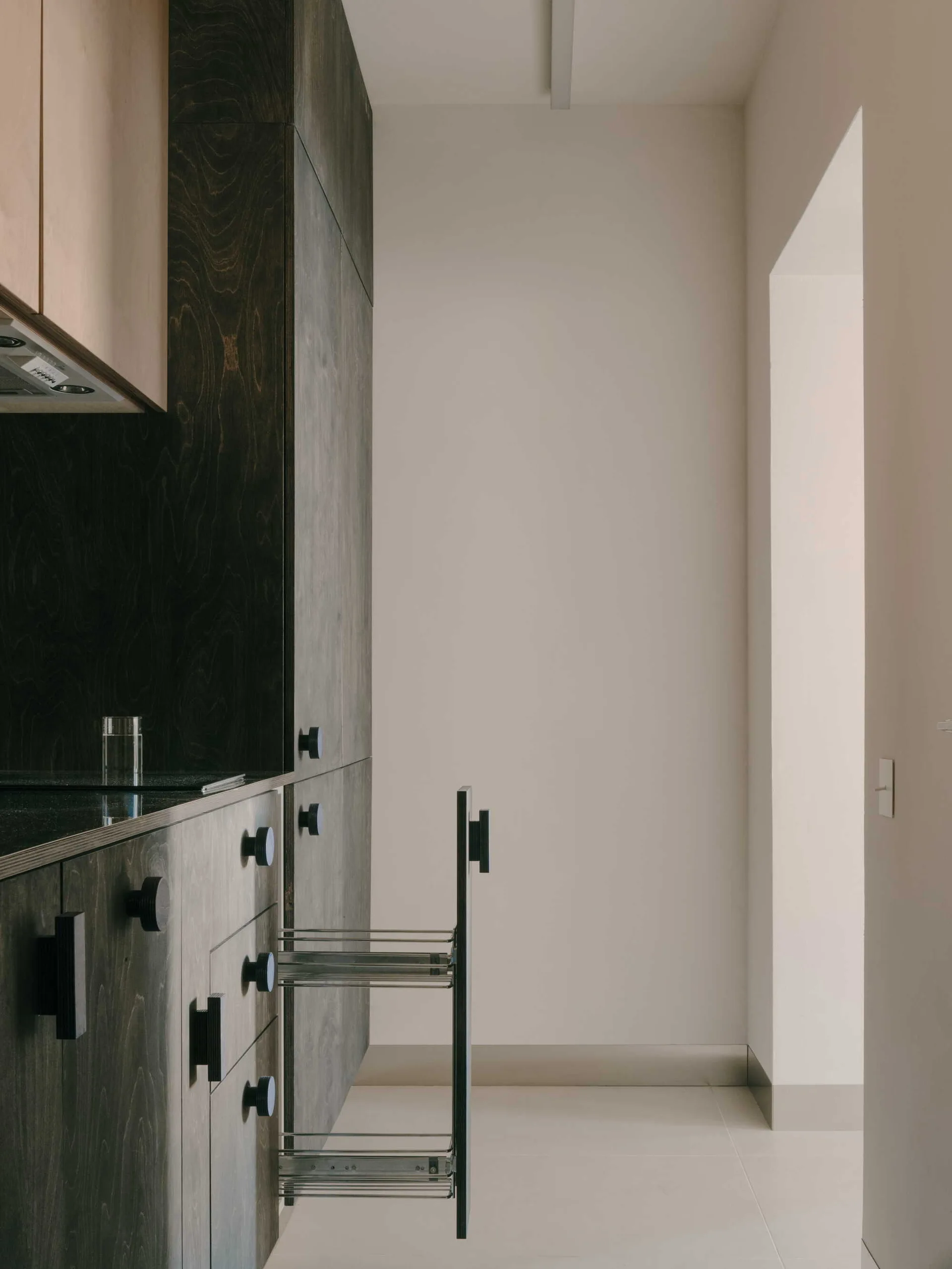 Minimalist dark wood grain kitchen cabinets featuring integrated black knobs and an open metal pull-out pantry drawer next to an off-white wall.