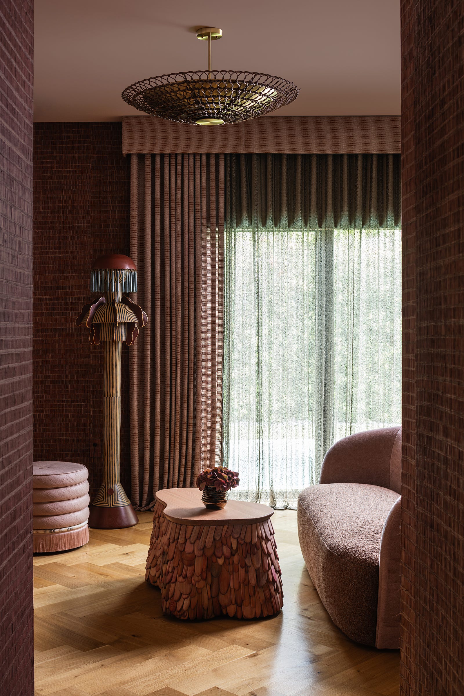 An eclectic seating corner features a curved dusty pink sofa, a textured wooden coffee table, a unique fringed floor lamp, and brown woven wallpaper.