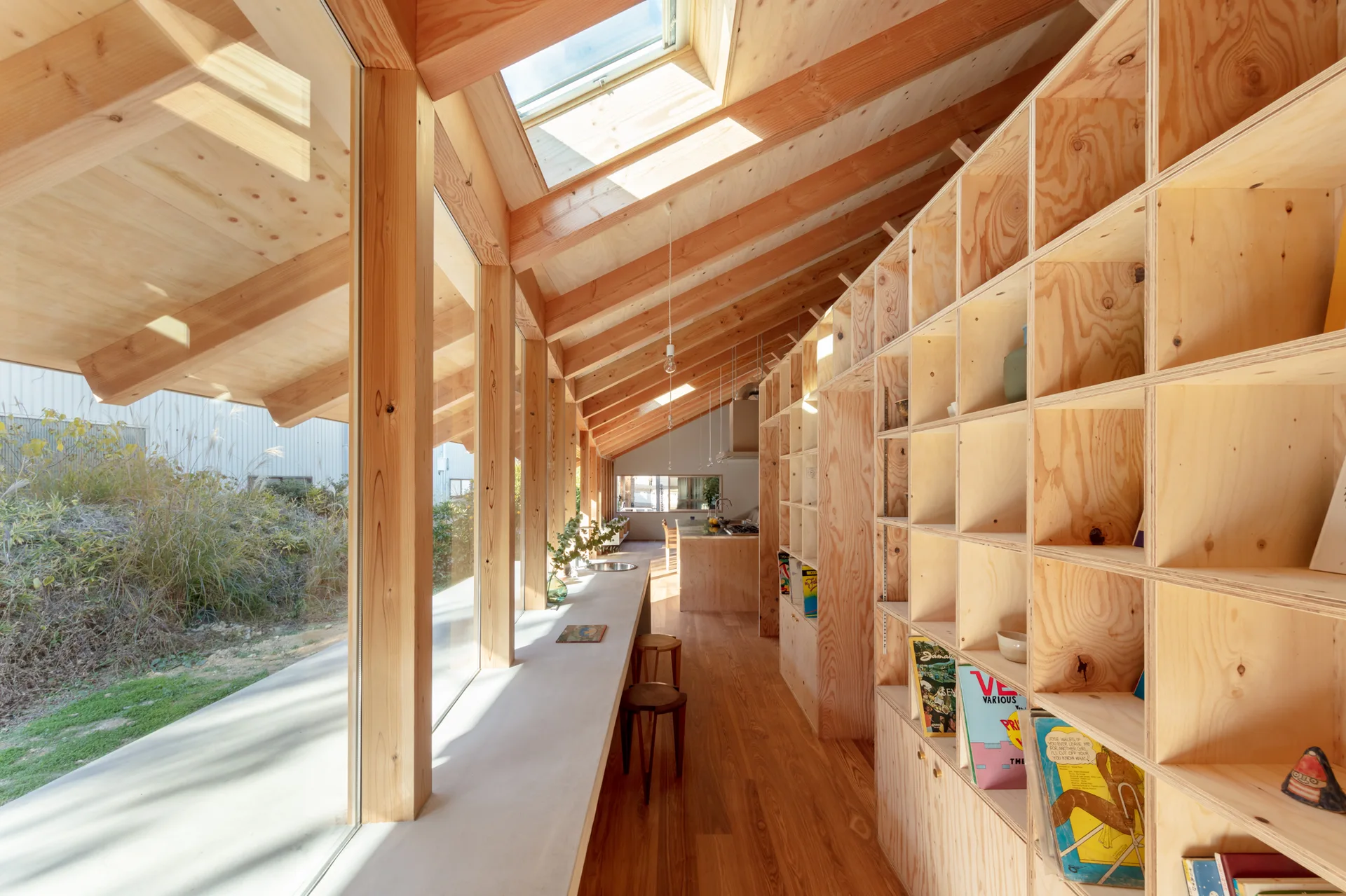 A bright interior space with exposed timber rafters, a long wall of plywood grid shelving, a concrete counter along large windows, and a skylight.