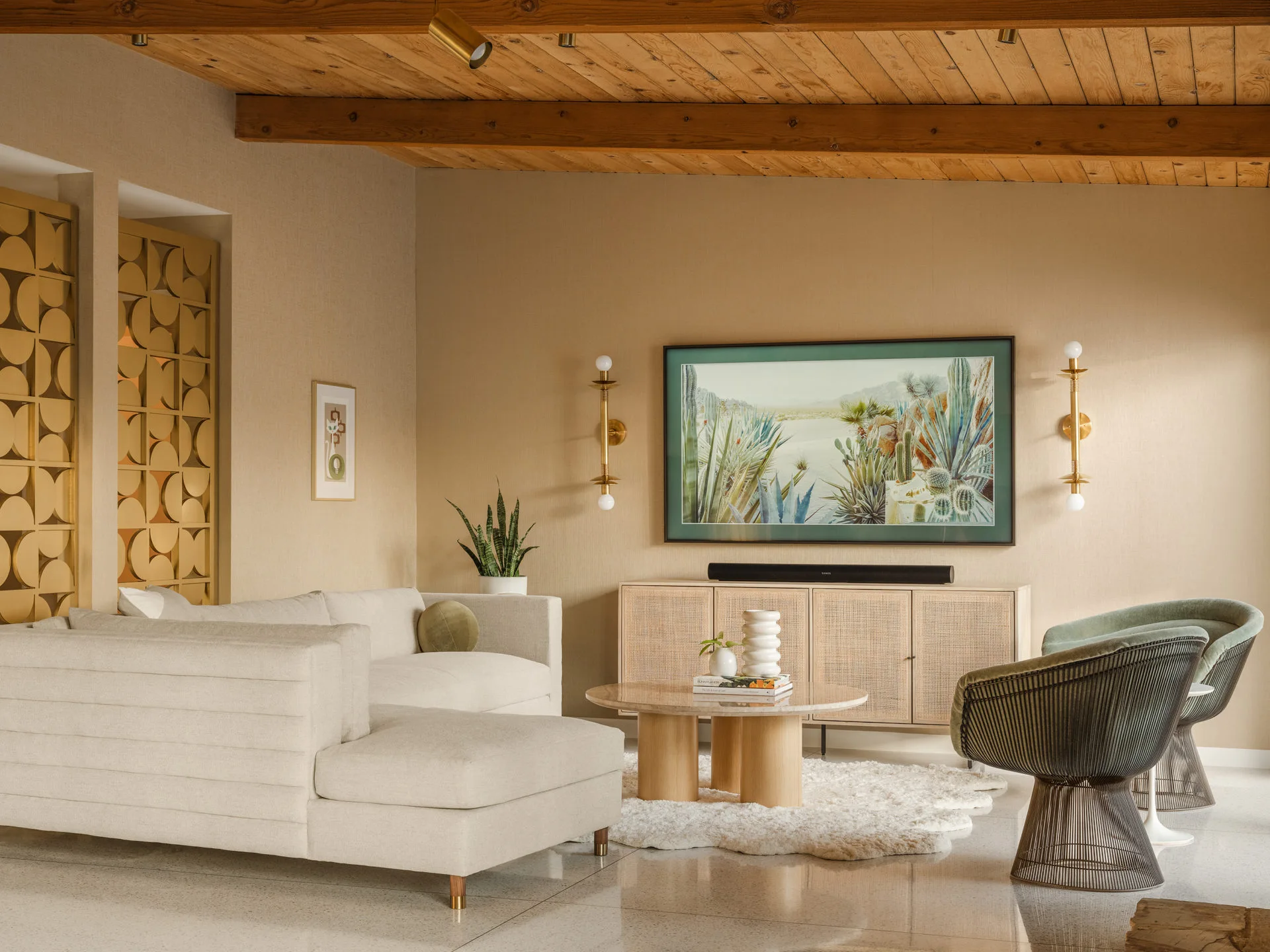 A modern living room features a cream sectional, caned media console, wood ceiling beams, and framed desert artwork on the wall.