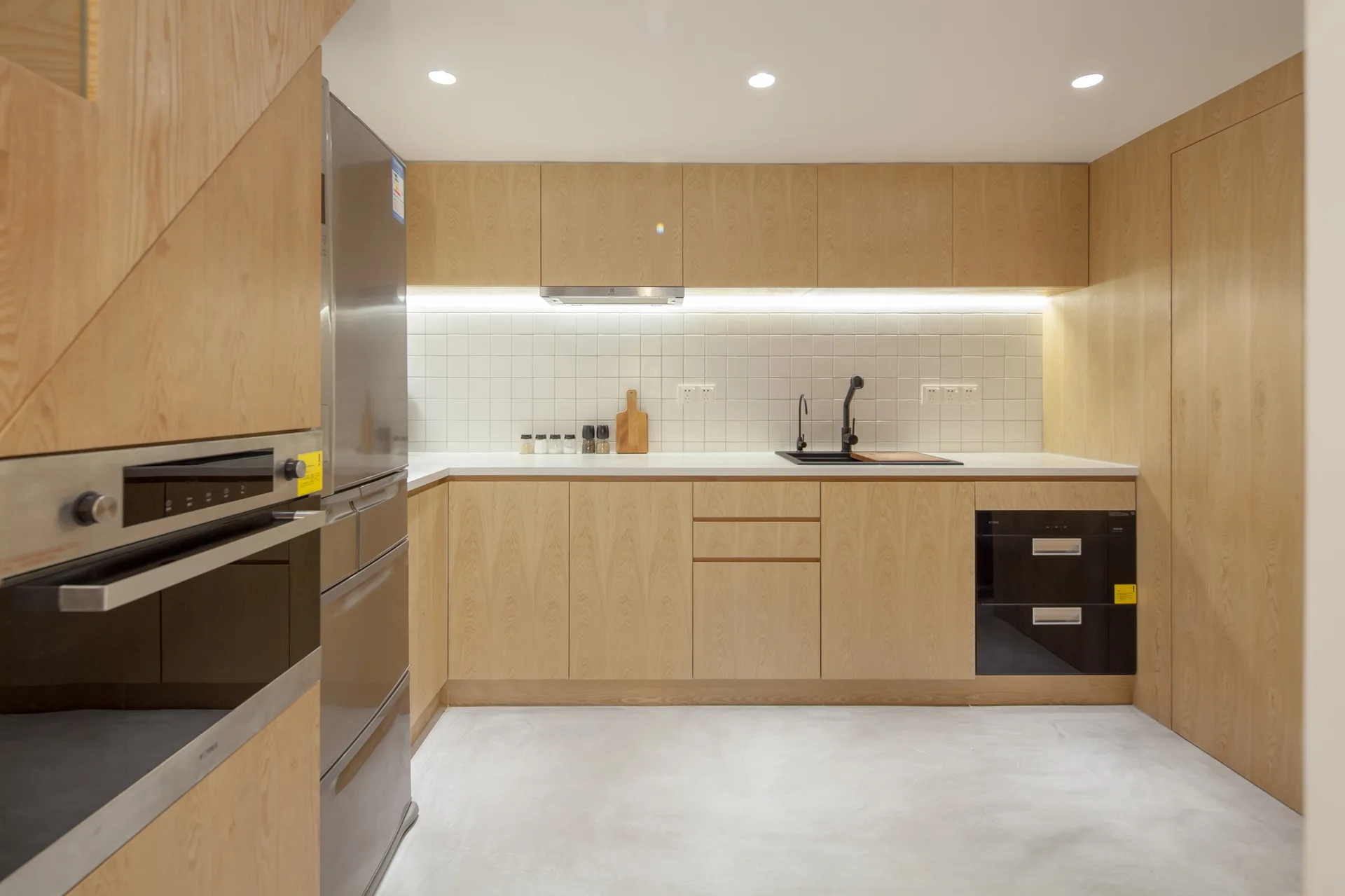 A minimalist kitchen features flat-front light wood cabinetry, a white square tile backsplash, and a clean concrete floor.