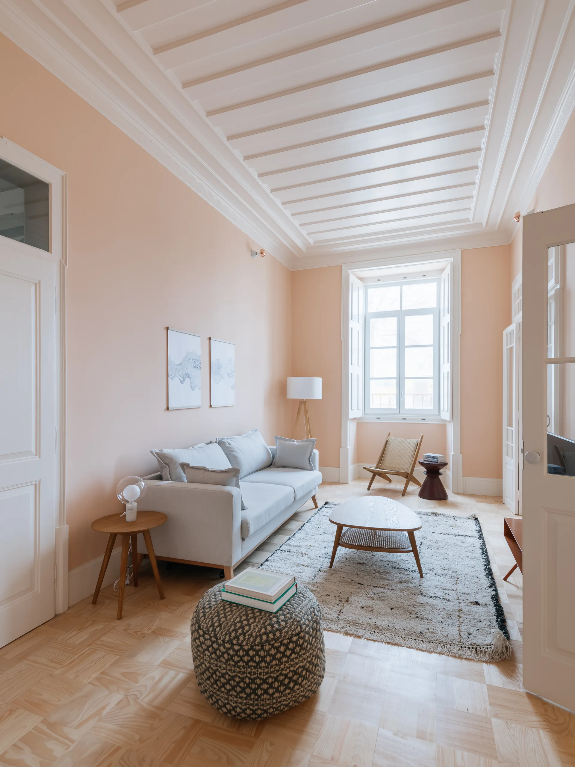 A Scandinavian living room with pale peach walls, a white wood paneled ceiling, a grey sofa, and light parquet flooring.