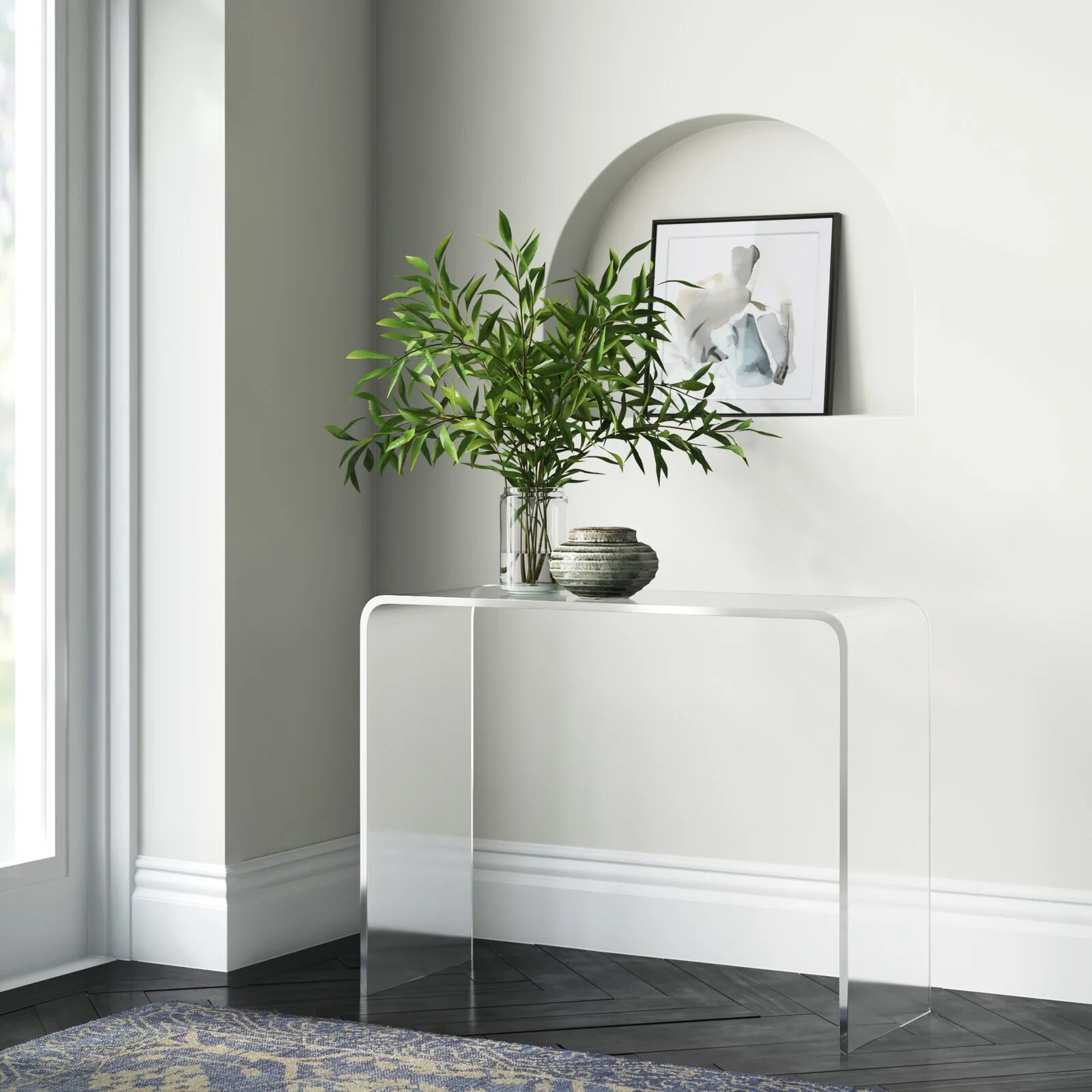 A clear acrylic waterfall console table holding greenery in a vase sits in front of a white wall with an arched alcove.