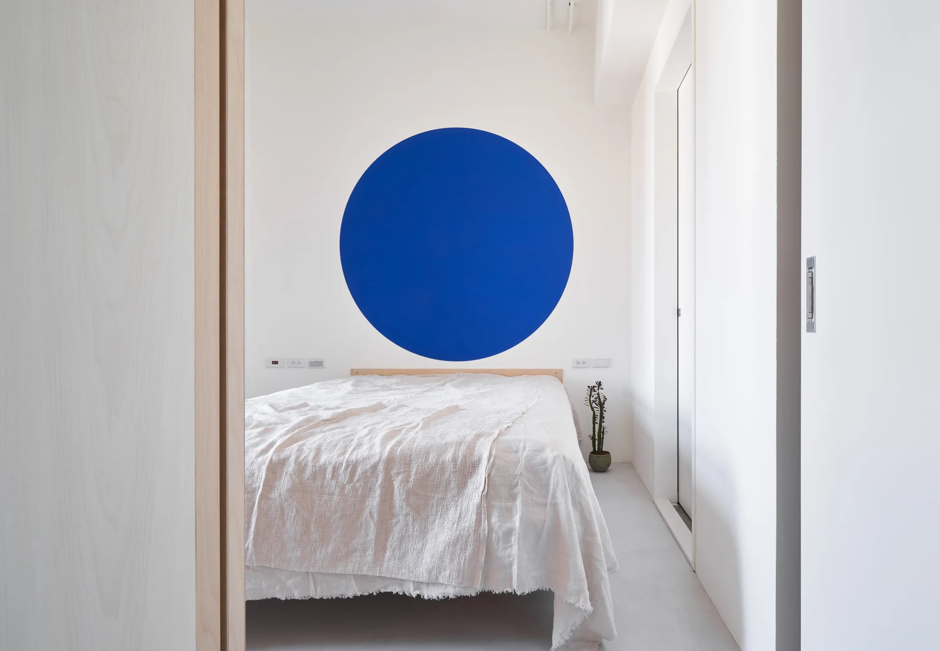 A minimalist bedroom featuring a low-profile bed with textured white linens in front of a white wall with a painted blue circle.
