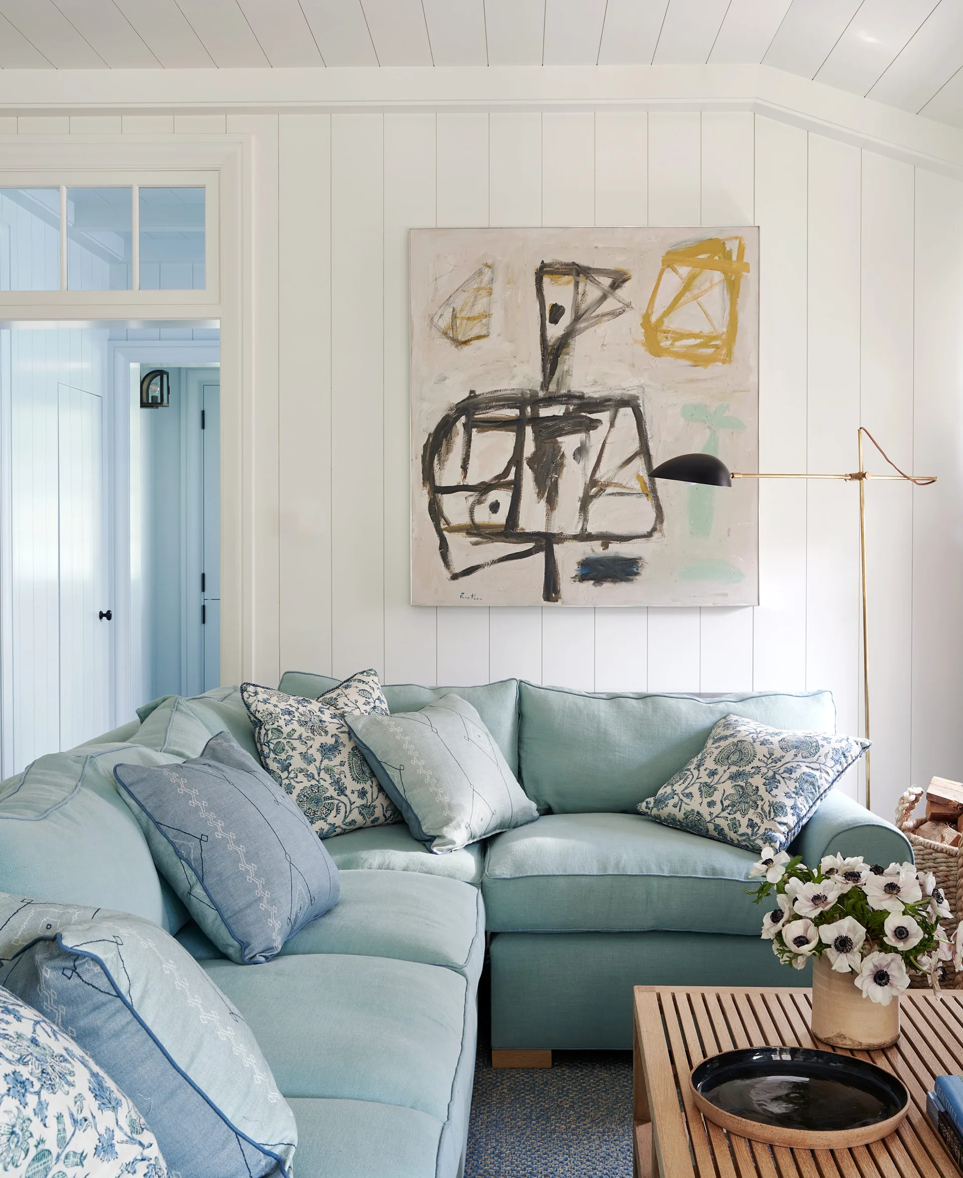 A light blue sectional sofa is paired with a wooden coffee table in a living room with white shiplap walls and abstract art.