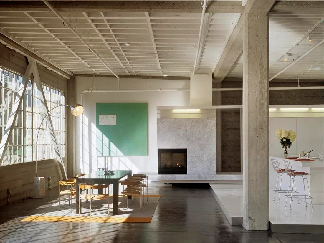 An industrial open-plan loft featuring a dining table, a marble fireplace, exposed concrete columns, and large grid windows letting in natural sunlight.