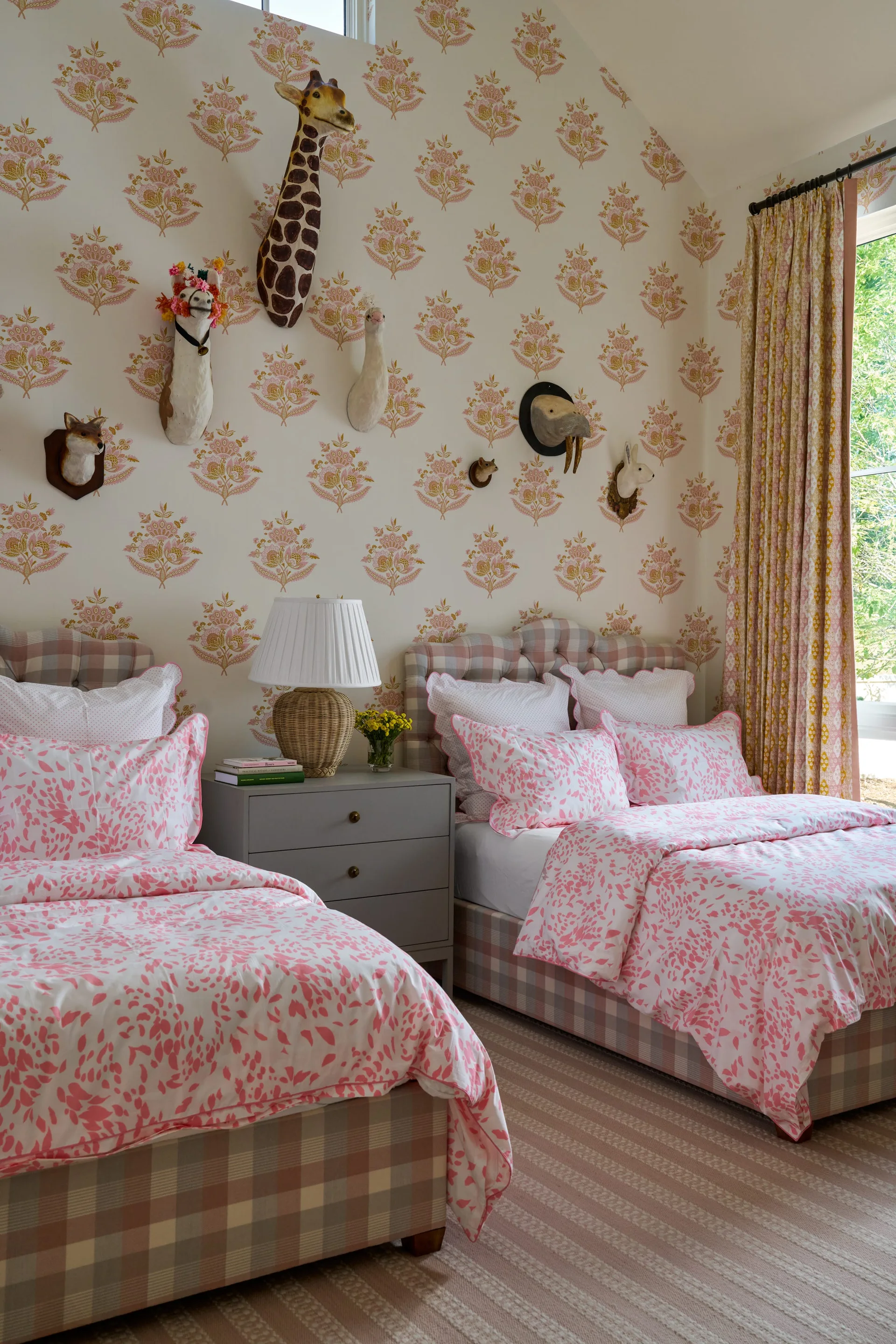 A children's bedroom features two twin beds with pink botanical bedding, a grey nightstand, and decorative animal busts mounted on floral wallpaper.