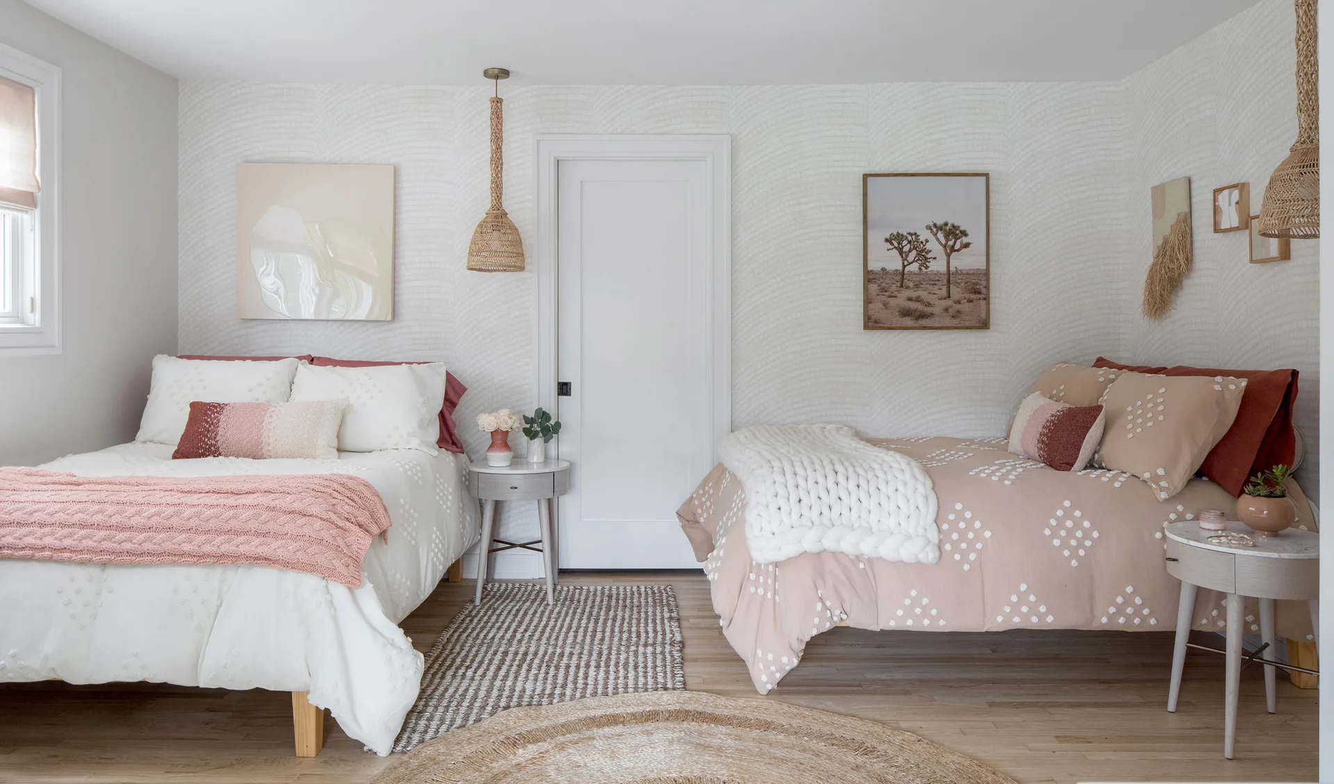 Two beds dressed in textured pink and white bedding flank a white interior door in a bedroom with patterned wallpaper and woven pendant lights.