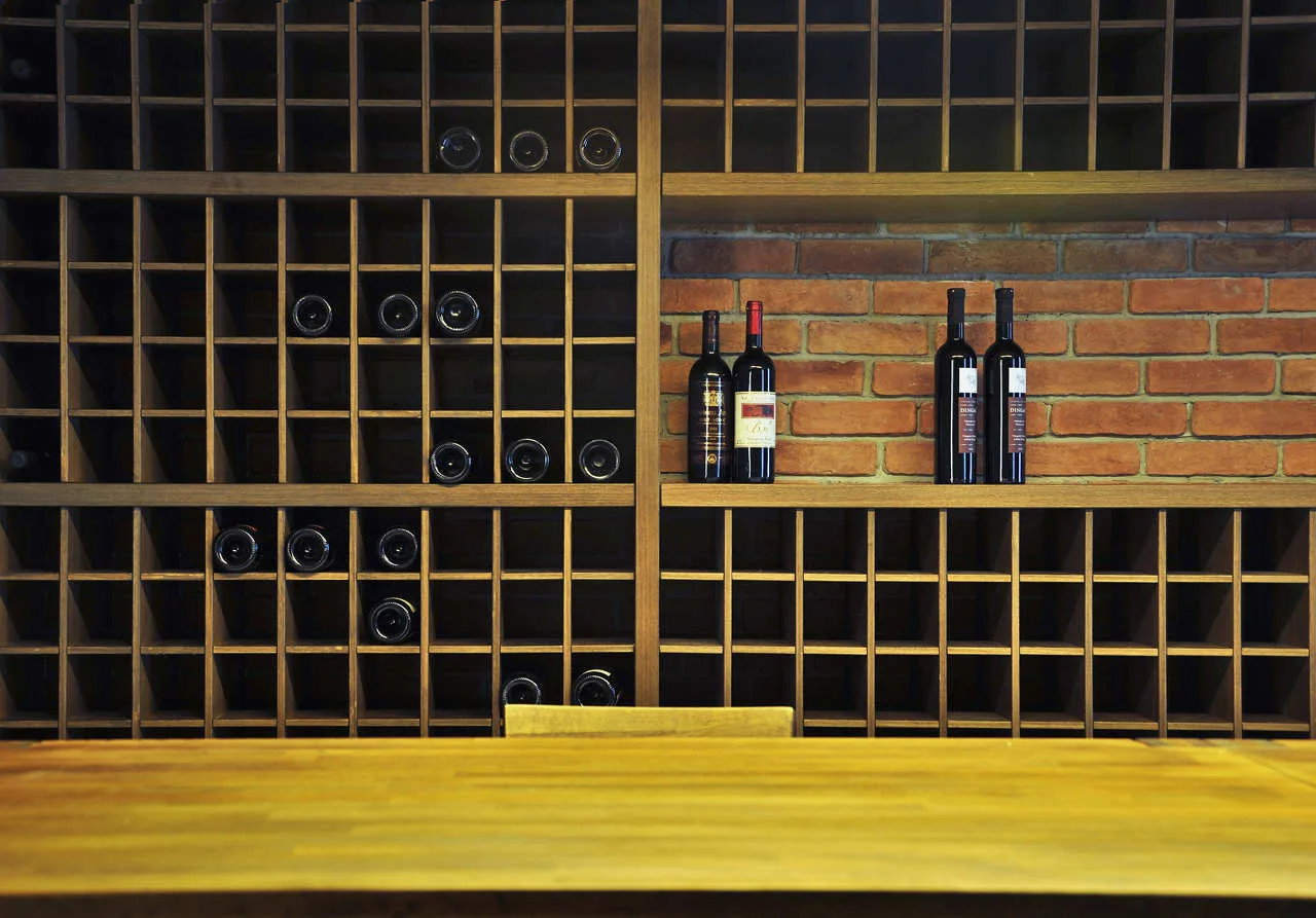 A dark wood grid wine rack is mounted against an exposed red brick wall with a wooden table surface in the foreground.